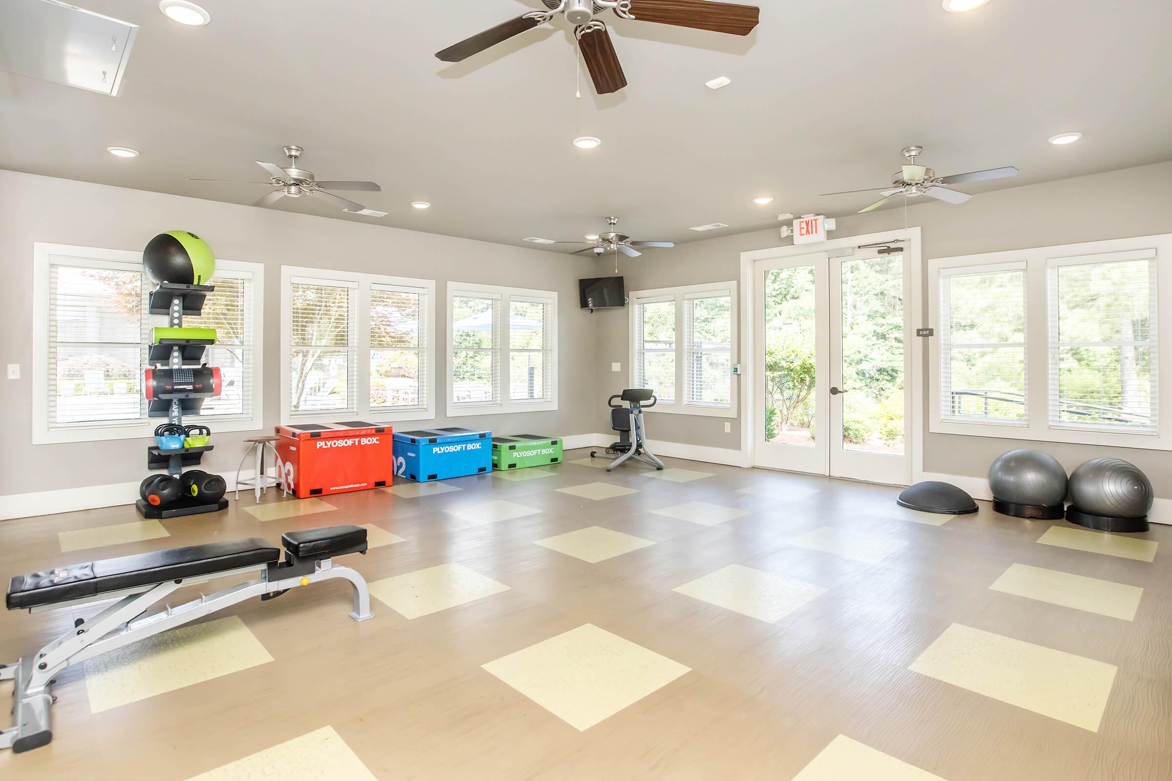 A bright, spacious fitness room with large windows, wooden floors, and a variety of exercise equipment. The room features a bench press, colorful weight storage bins, two stability balls, and ceiling fans for ventilation. Natural light fills the space, enhancing the open, inviting atmosphere.