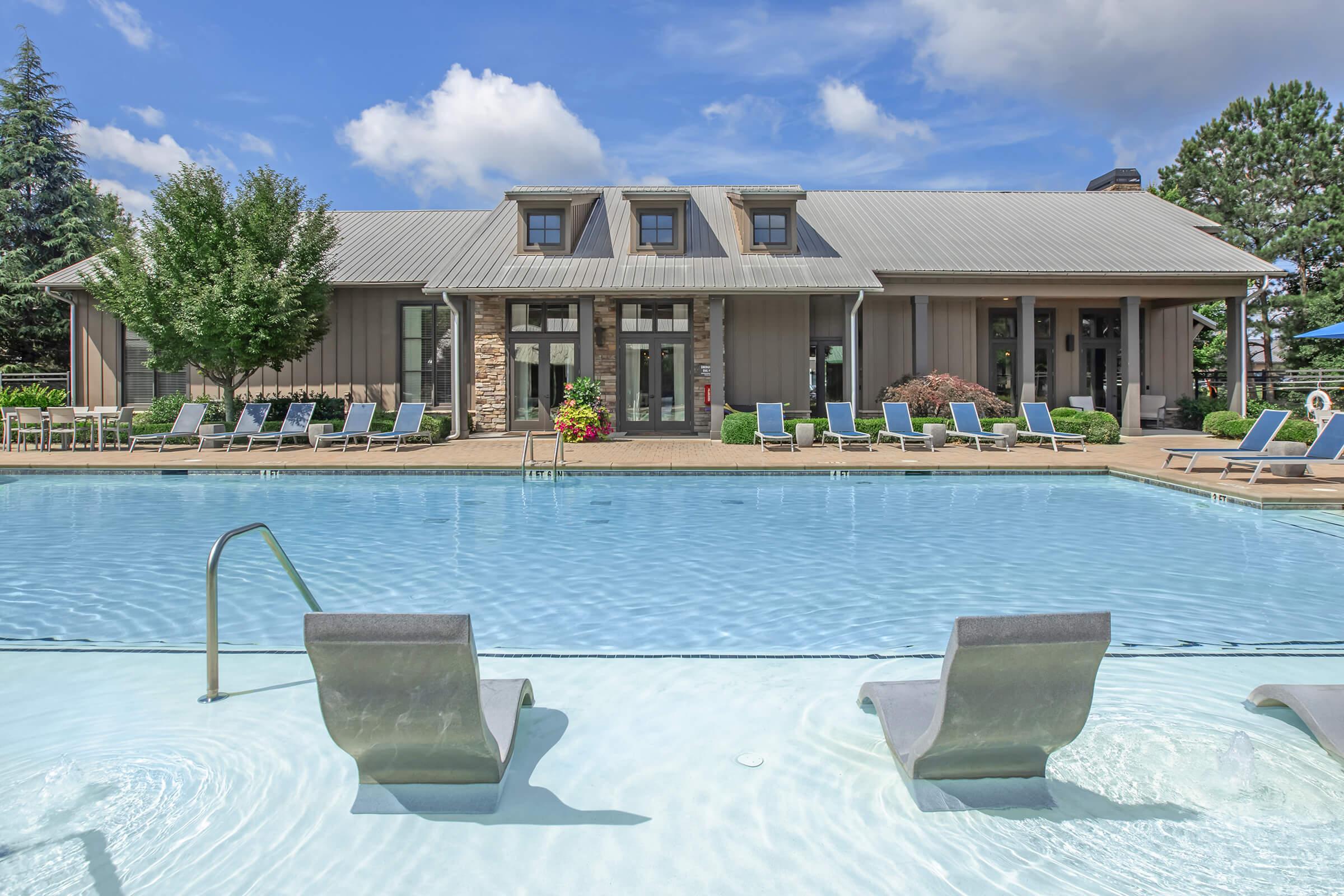 A bright blue swimming pool with two spa-like lounge chairs partially submerged in the water. In the background, a modern building with large windows is surrounded by trees and vibrant landscaping. The sky is clear with a few fluffy clouds, creating a sunny and inviting atmosphere.