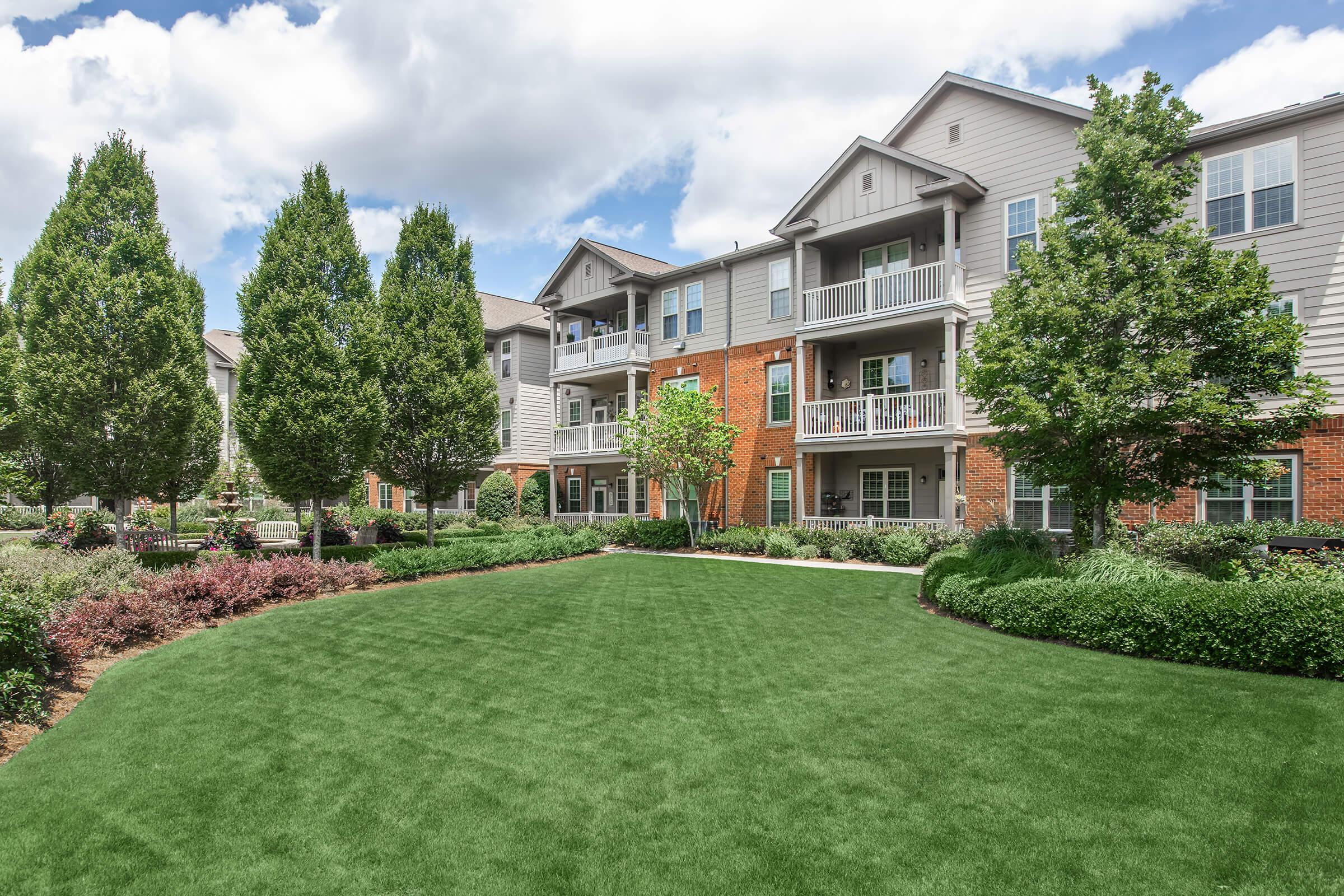 A well-maintained apartment complex featuring multiple buildings, lush green lawns, and landscaped areas with shrubs and trees. The scene is set against a backdrop of partly cloudy skies, showcasing a serene residential environment.
