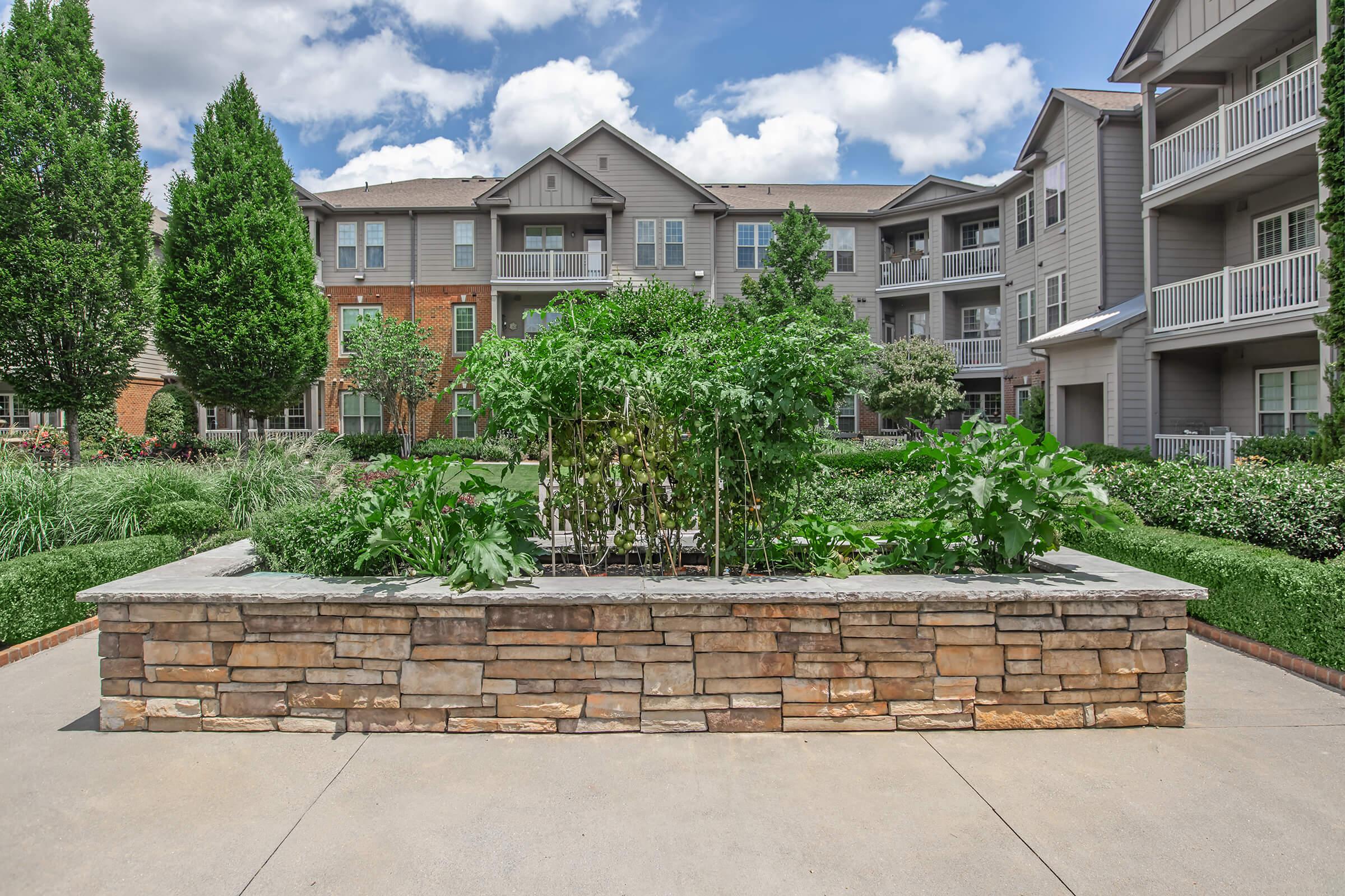 A landscaped courtyard featuring a stone planter filled with lush green plants and vegetables. Surrounding the planter are trimmed hedges and trees, with several apartment buildings in the background under a partly cloudy sky. The scene conveys a serene and inviting outdoor living space.