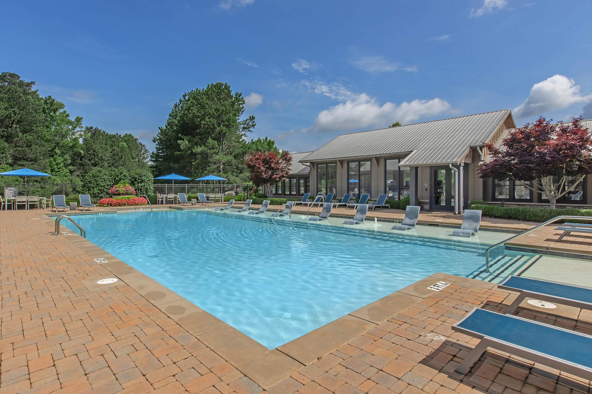 A serene outdoor pool area featuring a crystal-clear pool with lounge chairs and umbrellas. Surrounding the pool are landscaped gardens with trees and flowers, and a clubhouse in the background. The sky is bright blue with a few fluffy clouds, creating a relaxing atmosphere.