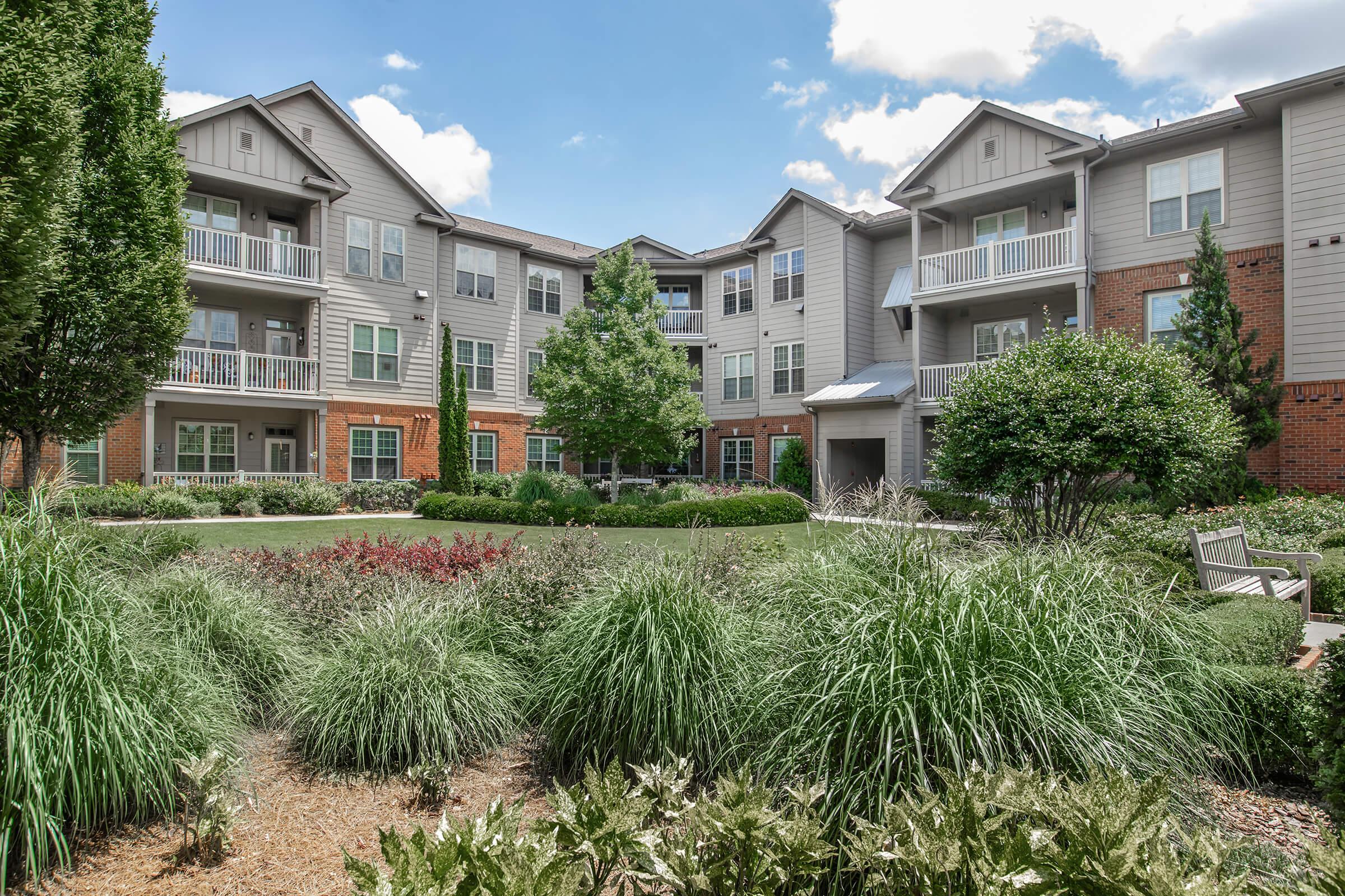 A well-maintained apartment complex courtyard featuring lush green shrubs, ornamental grasses, and flowering plants. The buildings in the background have multiple balconies and large windows, surrounded by trees under a blue sky with fluffy white clouds.