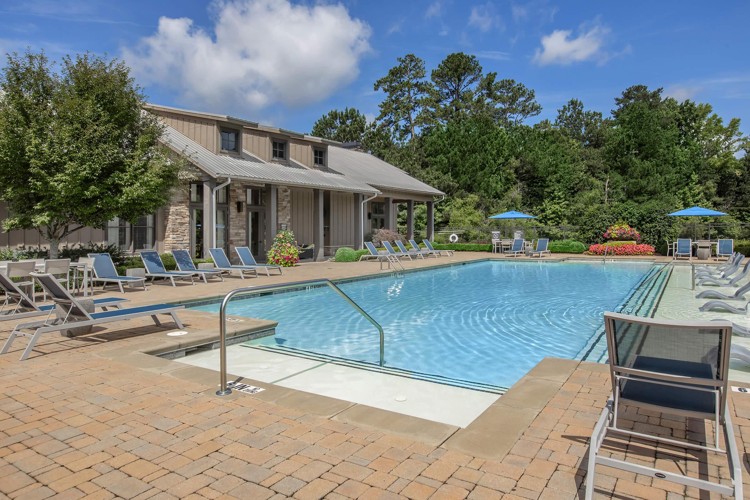 A sunny outdoor pool area featuring a large, clear blue swimming pool surrounded by lounge chairs. There's a building with a modern design in the background, along with trees and colorful flower beds. Umbrellas provide shade for the seating areas.