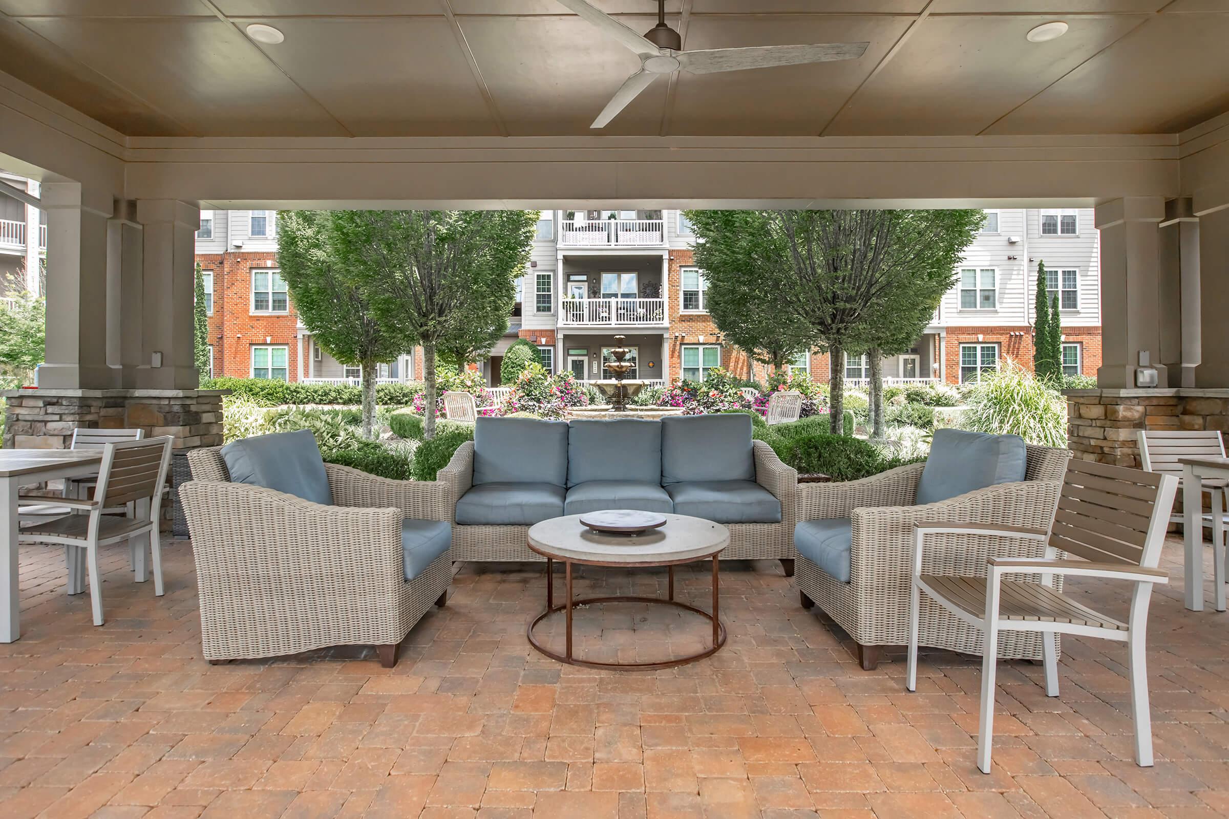 A shaded outdoor seating area featuring a light blue cushioned sofa and chairs surrounding a round coffee table. Lush greenery and flowering plants are visible in the background, enhancing the inviting atmosphere of the space.