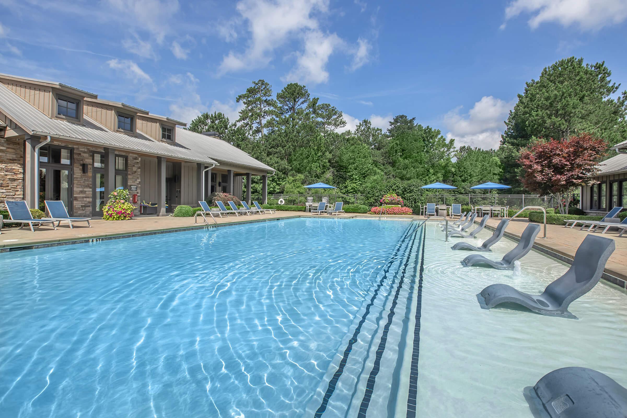 A serene outdoor swimming pool surrounded by lounge chairs and umbrellas, with a backdrop of lush trees and a building. The crystal-clear water reflects the blue sky and fluffy clouds, creating a tranquil and inviting atmosphere for relaxation.