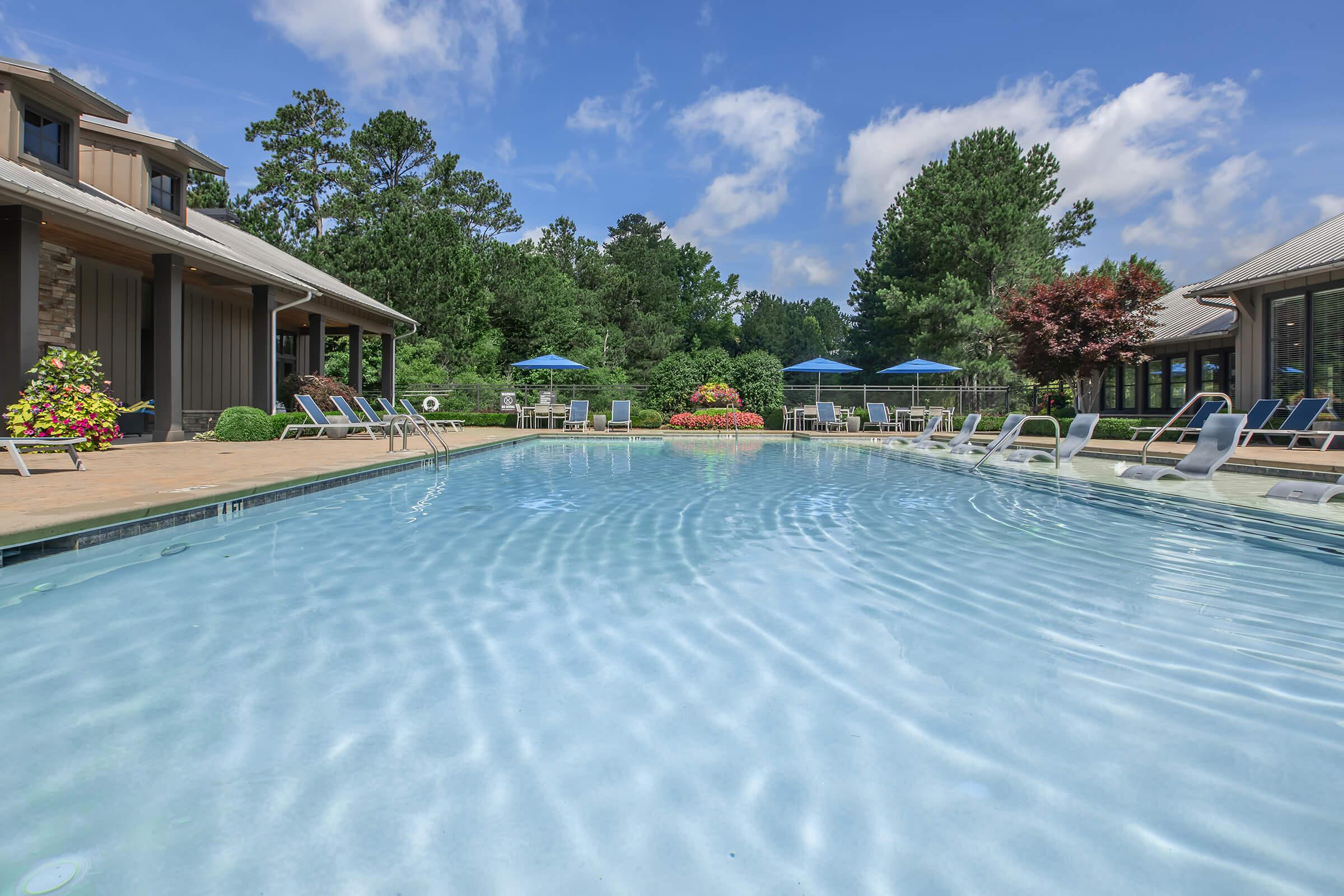A serene swimming pool surrounded by lounge chairs and colorful flower beds. The scene is framed by lush greenery and trees under a bright blue sky with scattered clouds. Sun umbrellas provide shade, creating a relaxing atmosphere for leisure and enjoyment.