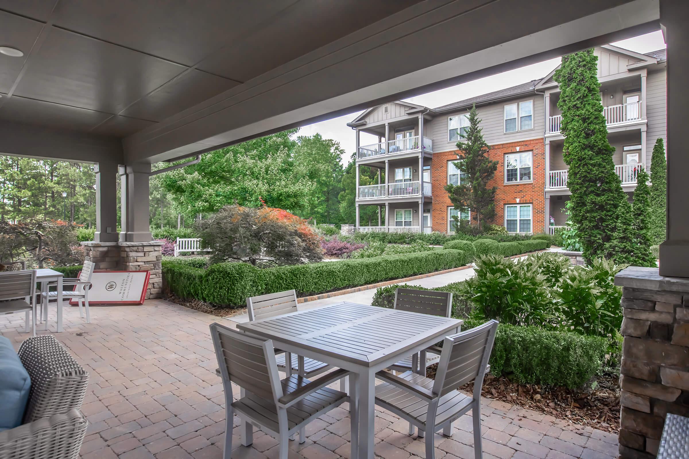 A covered patio area with a table and chairs overlooks a landscaped garden featuring shrubs and trees. In the background, there is a multi-story apartment building with balconies. The scene is calm and designed for relaxation, surrounded by greenery.