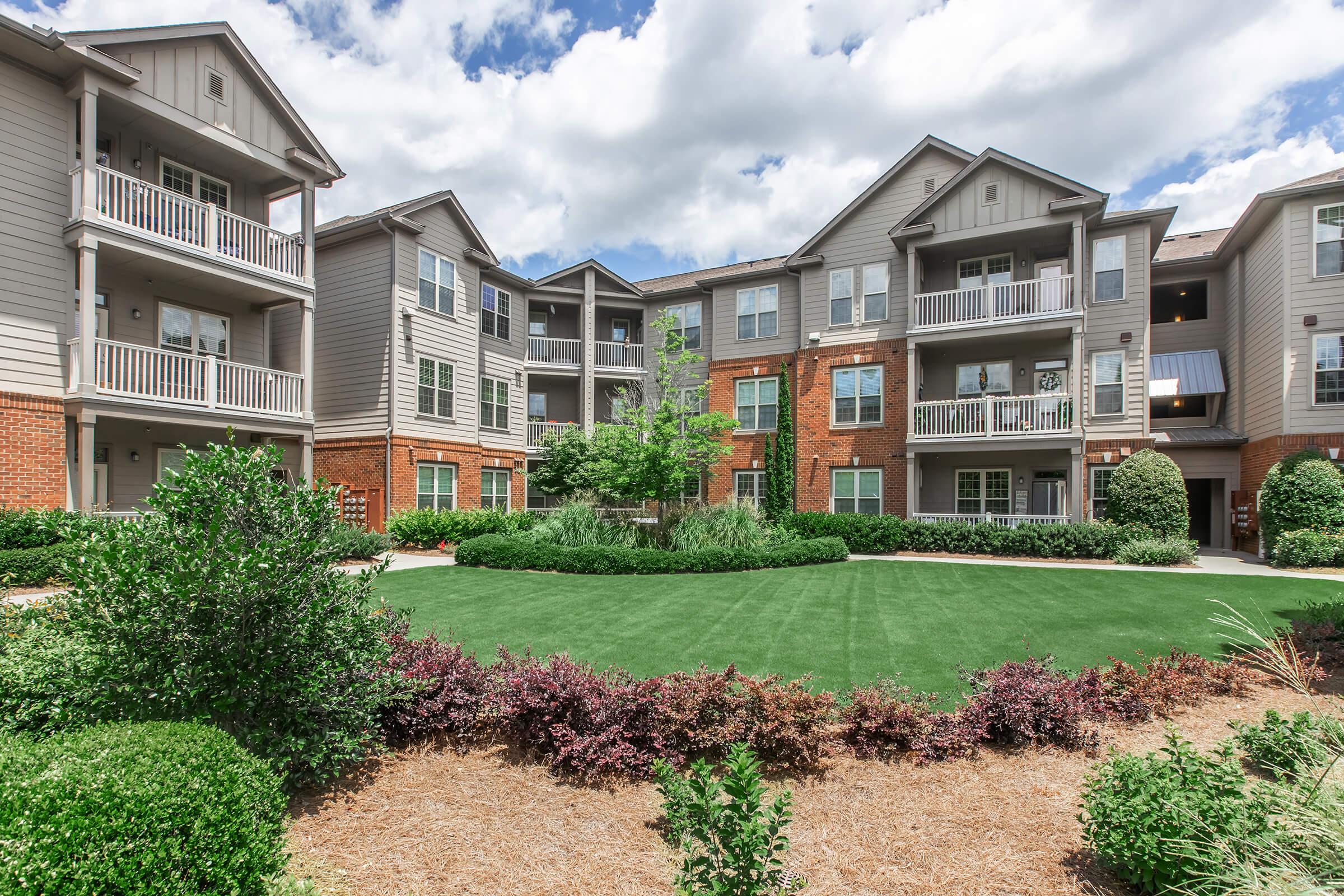 A landscaped courtyard featuring well-maintained grass and shrubs, surrounded by multi-story apartment buildings with balconies. The sky is partly cloudy, adding a bright atmosphere to the scene.