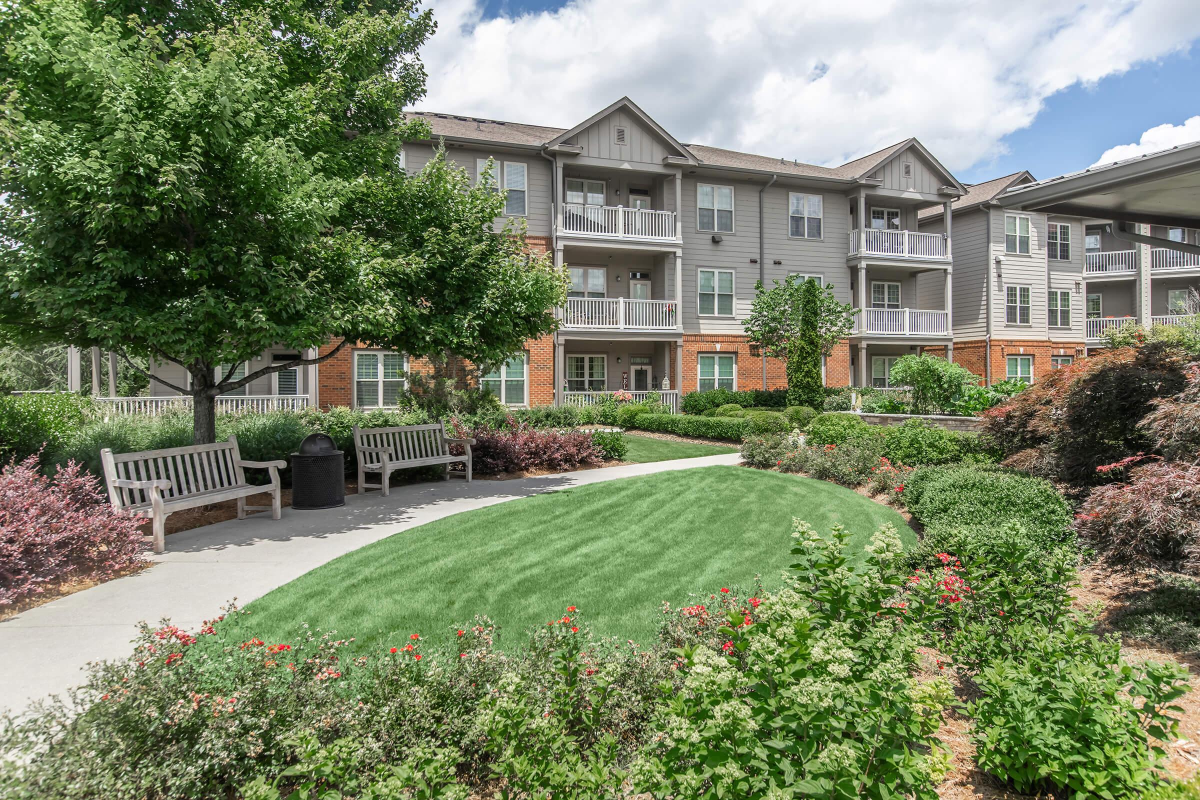 A well-maintained landscaped area featuring green grass, blooming flower beds, and benches. In the background, there are multi-story residential buildings with balconies. The sky is partly cloudy, contributing to a pleasant outdoor atmosphere.