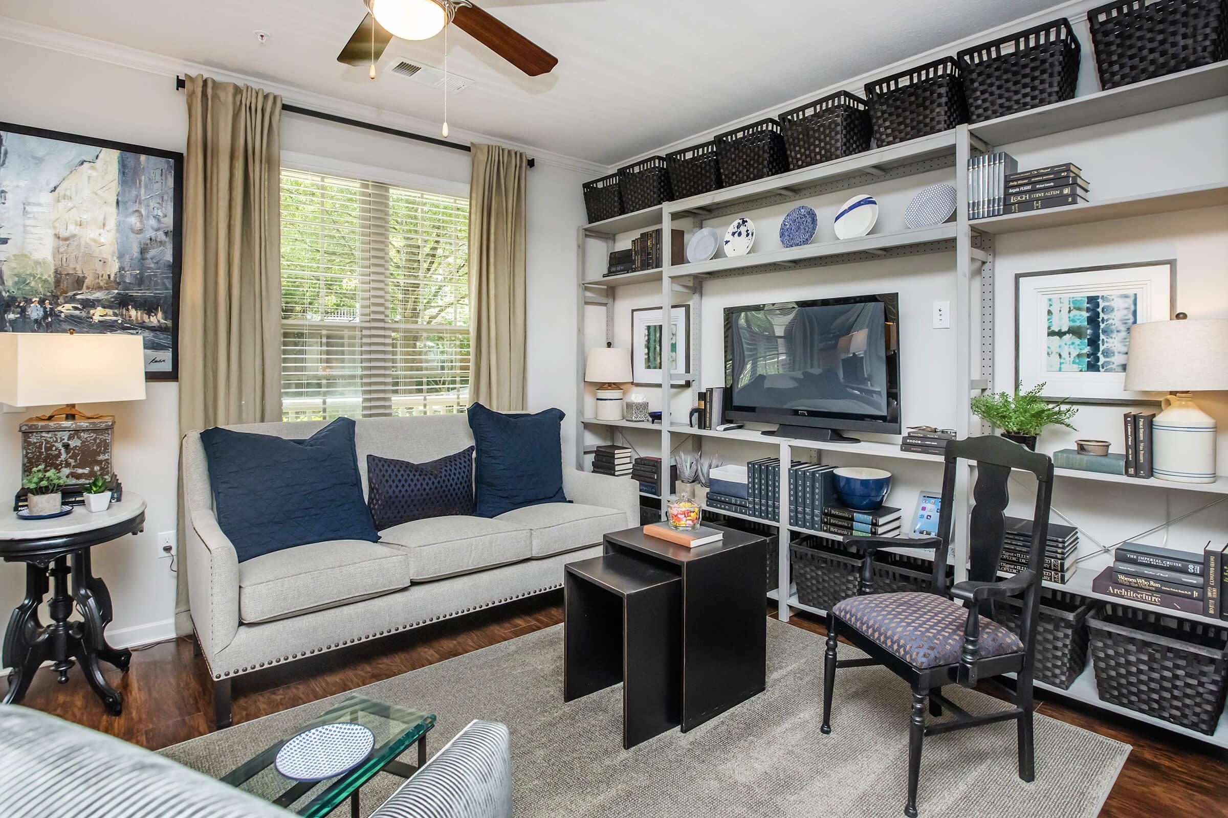 A cozy living room with a light-colored sofa adorned with dark blue cushions. It features a black coffee table, a shelf with neatly arranged books and decorative items, and a wall-mounted TV. Natural light filters through windows with curtains, creating a warm and inviting atmosphere.