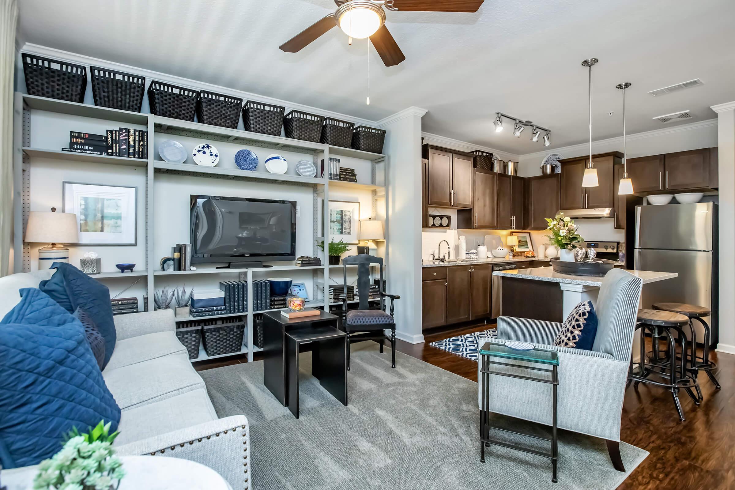 A cozy modern living room featuring a light gray sofa with blue accent pillows, a dark wood coffee table, and open shelving with decorative baskets. A flat-screen TV is mounted on the wall, with a kitchen area visible in the background, showcasing dark cabinets and stainless steel appliances.
