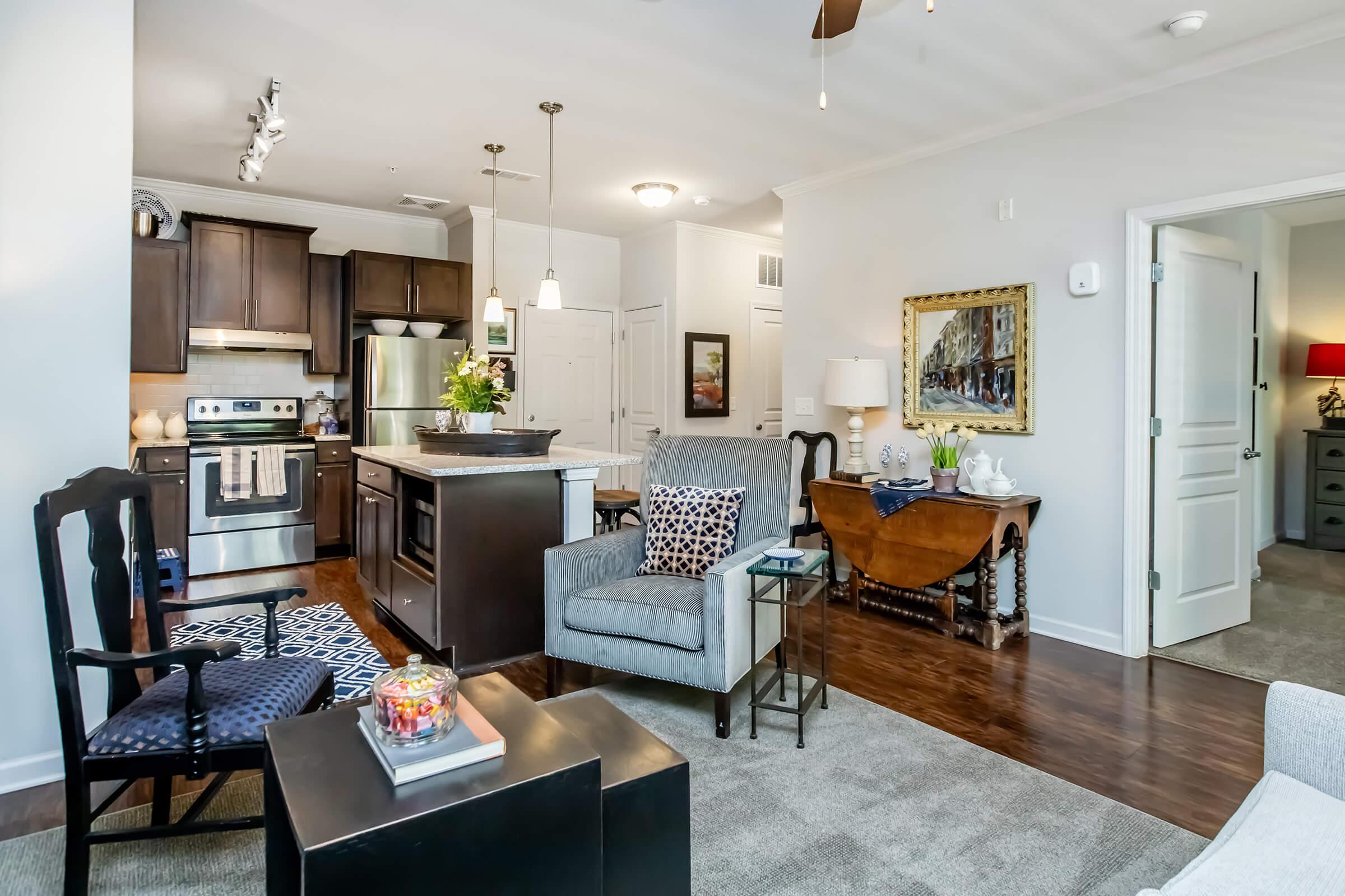 A modern living space featuring a kitchen with dark wood cabinets and stainless steel appliances. The area includes a cozy seating arrangement with a gray armchair, a patterned rug, and a small coffee table. A wooden dining table is visible, along with artwork on the walls, enhancing the inviting atmosphere.