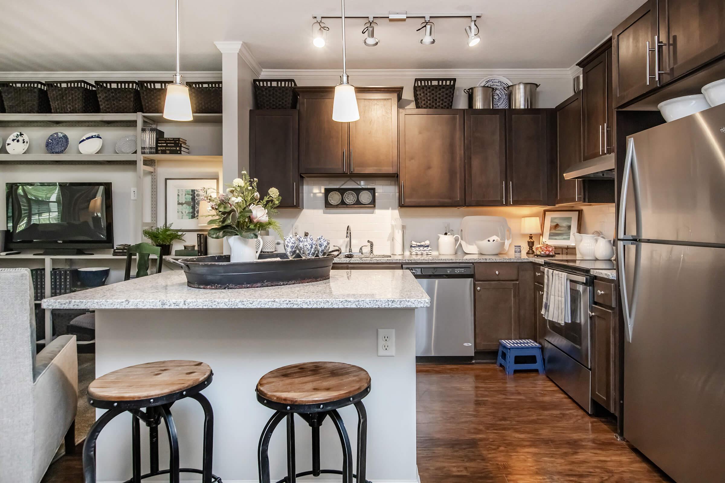 Modern kitchen with dark wooden cabinets, stainless steel appliances, and a large granite island. Two wooden stools are in front of the island. The decor includes potted plants and organized shelves with various dishes. Soft lighting from pendant lights creates a warm atmosphere.