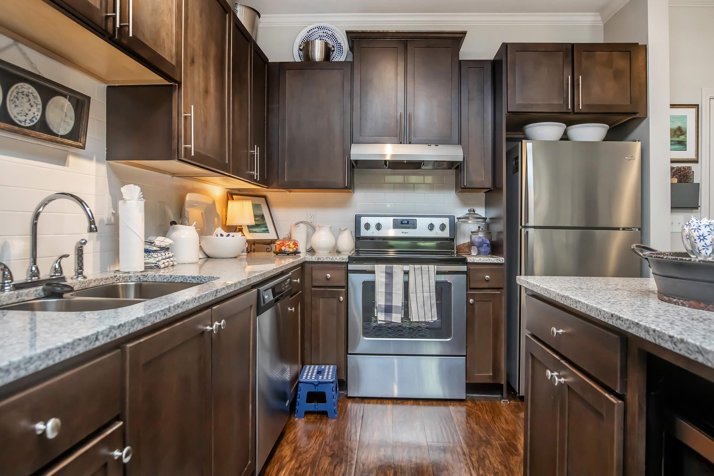 A modern kitchen featuring dark wood cabinets, a stainless steel refrigerator, and an oven with a sleek design. The countertop is granite, complemented by various kitchenware and decorative items. A small blue step stool is placed near the sink. Natural light brightens the space, giving it a cozy atmosphere.