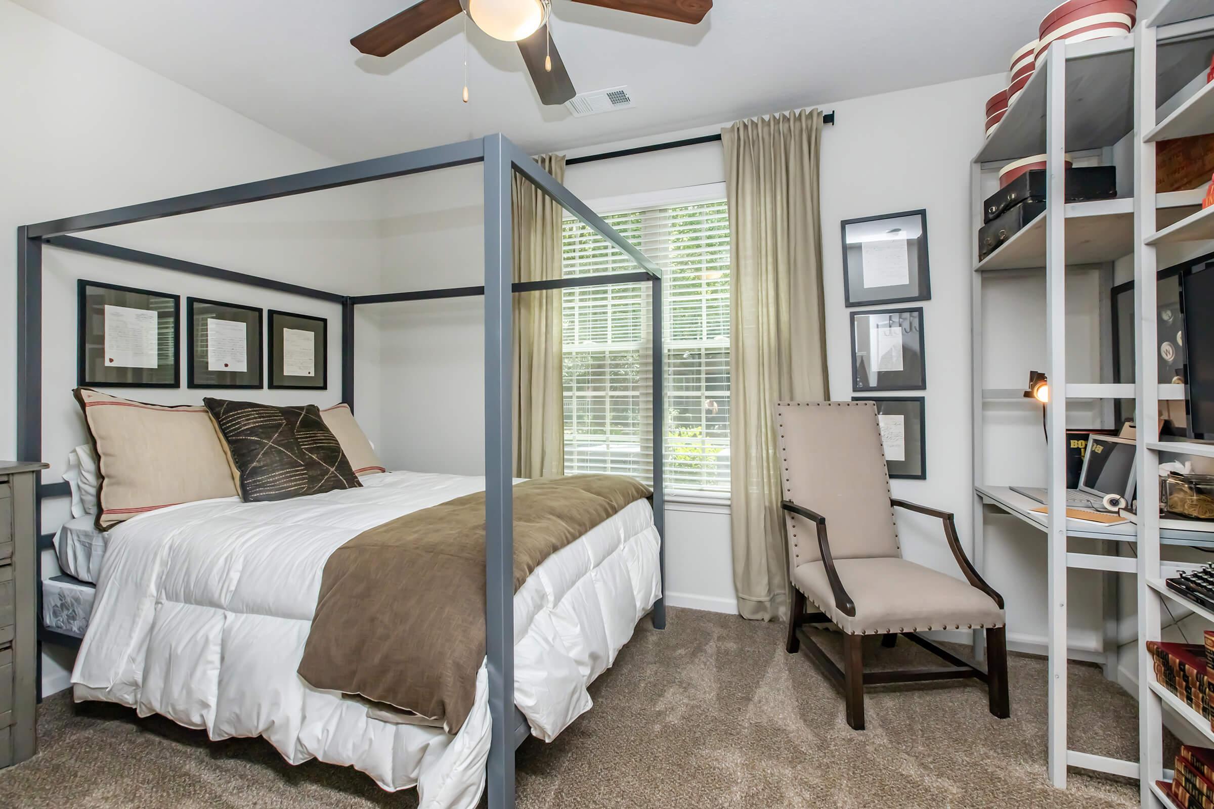 A cozy bedroom featuring a four-poster bed with white bedding and decorative pillows. There are framed pictures on the wall, a window with sheer curtains allowing natural light, and a stylish chair next to a bookshelf filled with items. The room has a warm, inviting atmosphere with neutral tones.