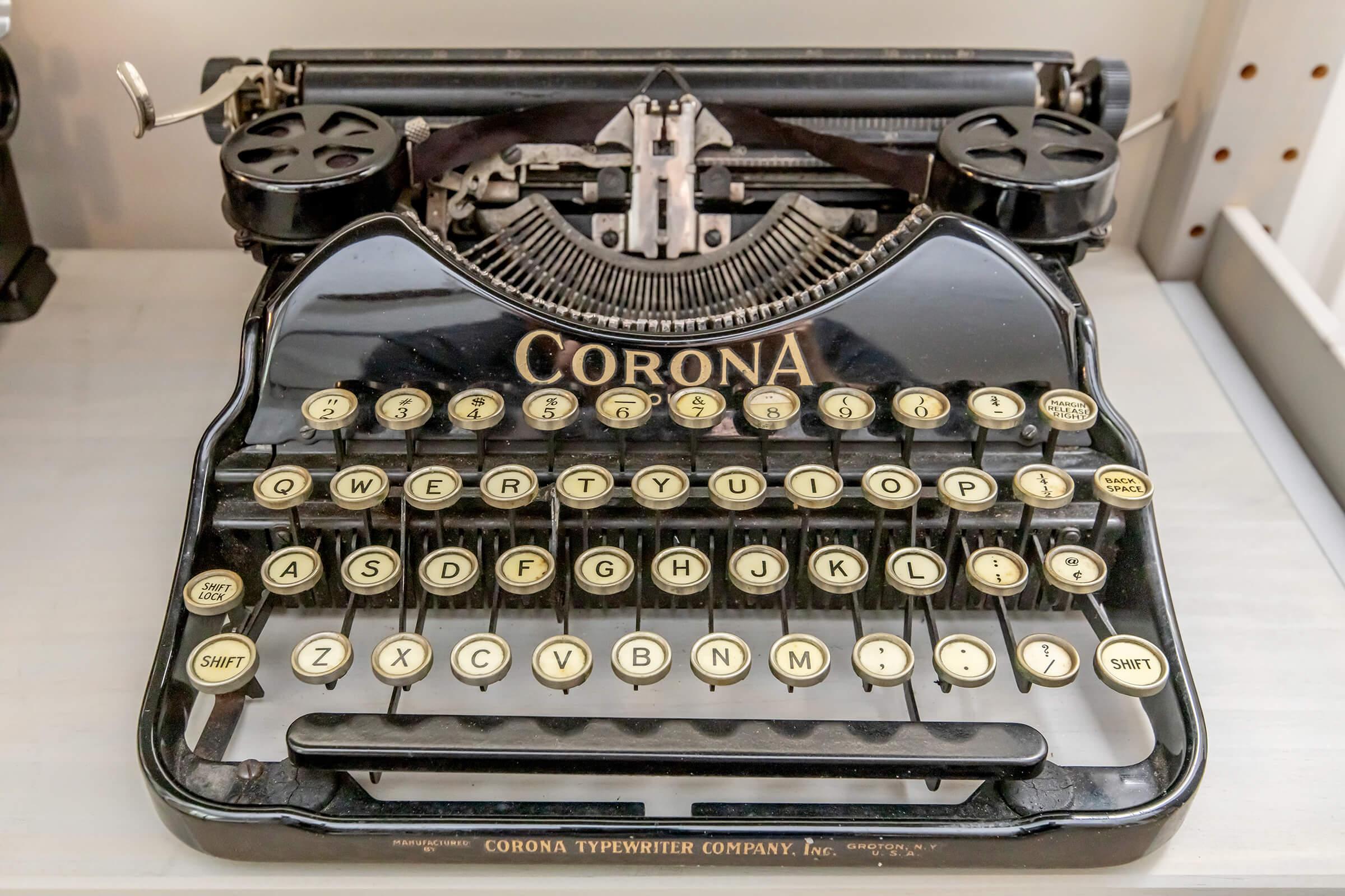 Antique Corona typewriter featuring a black body and round keys with white lettering. The typewriter has a classic design with a curved top panel and lever on the side, evoking a nostalgic era of writing technology. Designed for use on a smooth surface, it showcases craftsmanship from the early 20th century.