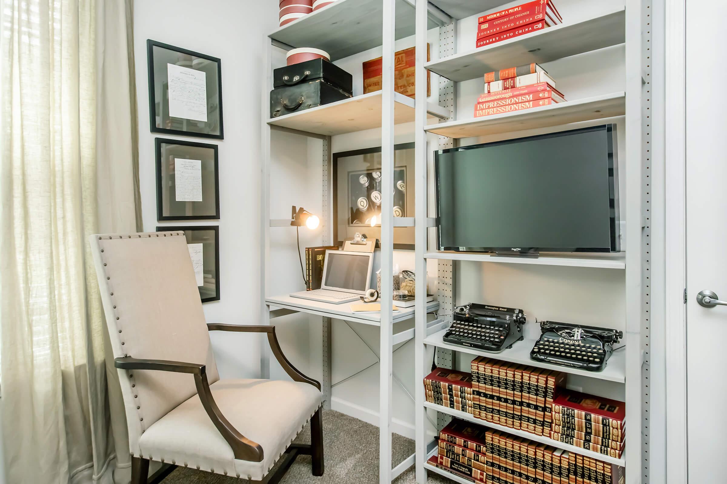 A cozy home office featuring a light-colored chair, a small desk with a laptop, and two vintage typewriters. Shelves hold books, framed documents, and decorative items. Natural light streams in through a nearby window, creating a warm and inviting workspace atmosphere.