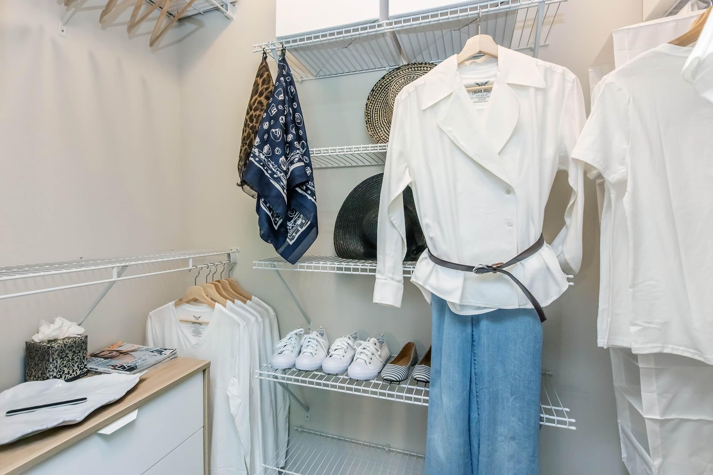 A neatly organized closet featuring a white blazer on a hanger, a pair of blue jeans, several white sneakers, and folded items. Accessories include scarves and decorative hats displayed on shelves, with a small table holding a tissue box. The overall aesthetic is minimalist and clean.