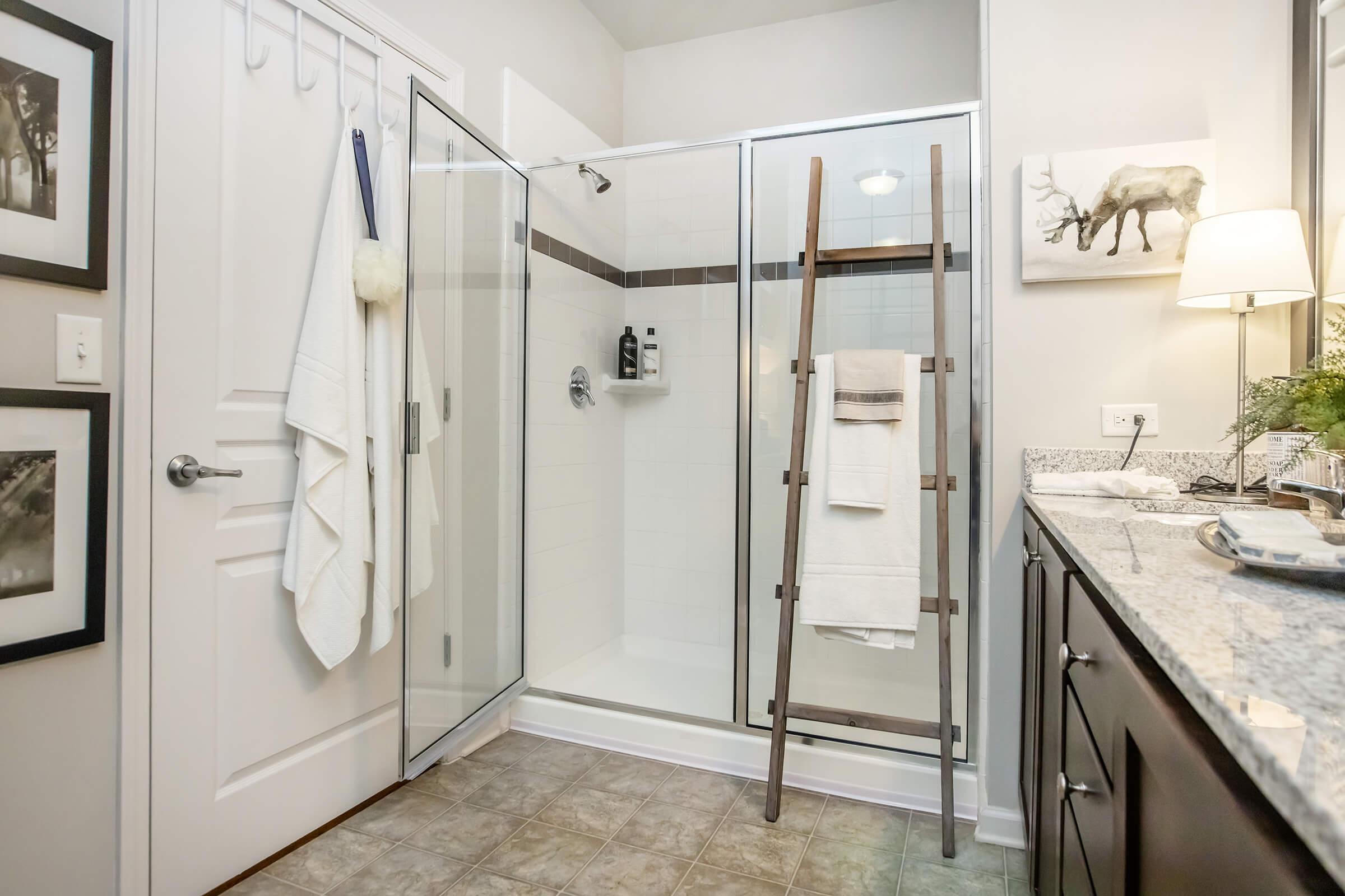 A modern bathroom featuring a glass shower enclosure, a wooden ladder holding towels, a stone countertop with toiletries, and framed black-and-white artwork on the walls. Soft lighting creates a warm atmosphere, complemented by neutral-colored tiles and walls.