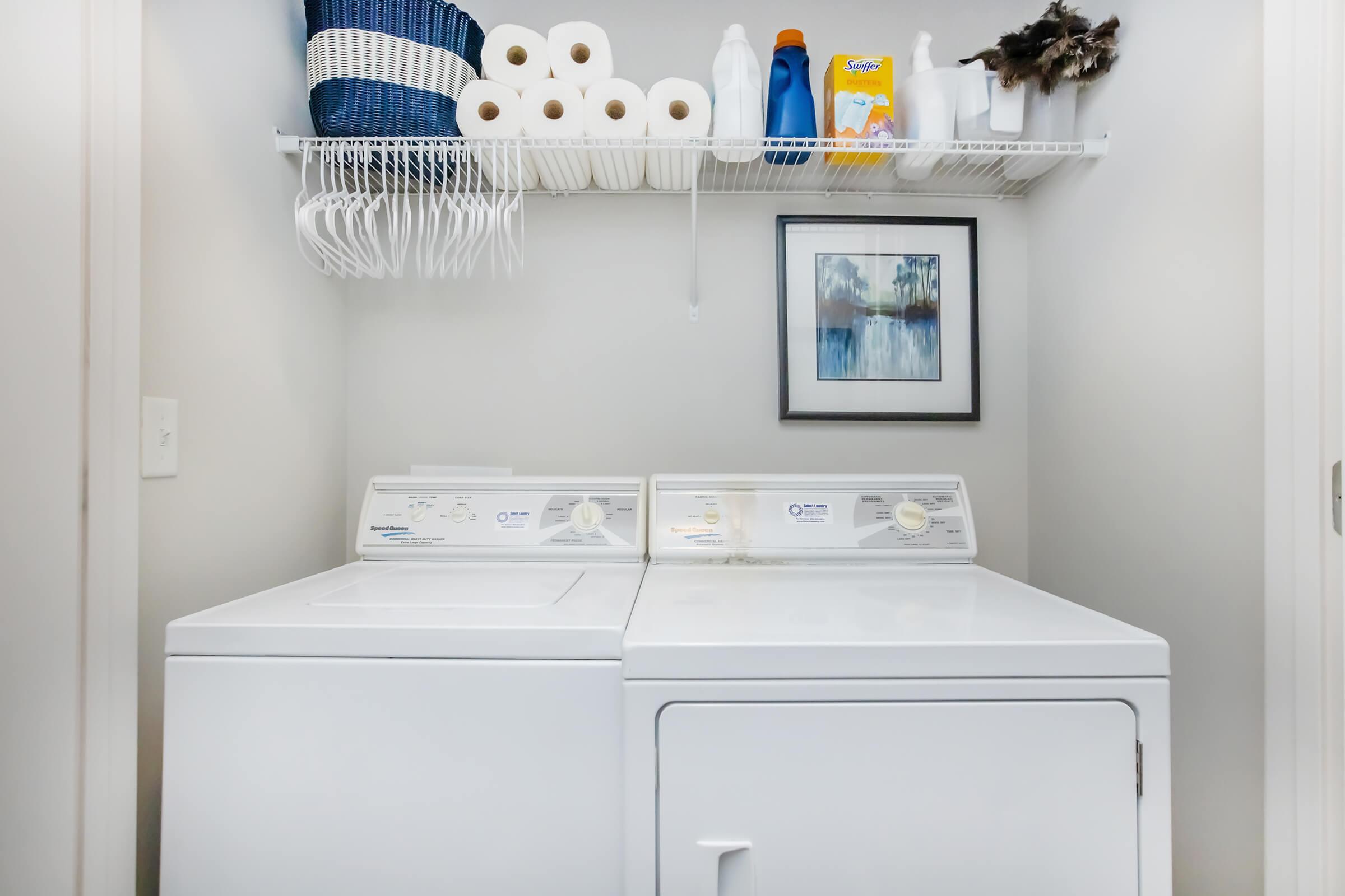 A laundry room featuring a white washer and dryer side by side. Above them, a shelf holds a blue basket, rolls of paper towels, cleaning supplies, and a framed artwork. The wall is painted a light color, creating a clean and organized space.