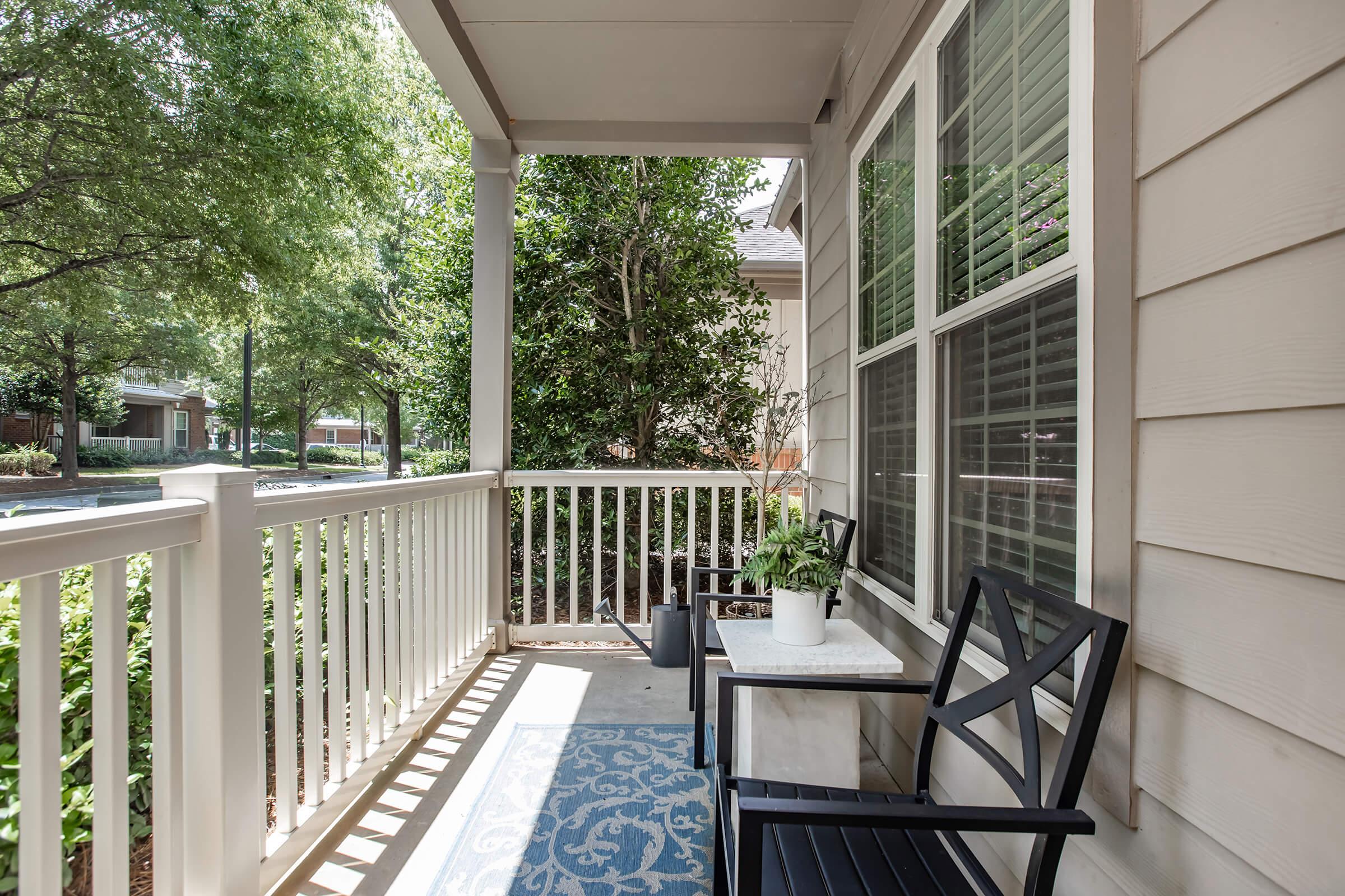 A cozy porch features two black chairs and a small table with a potted plant. Sunlight filters through leafy trees, casting shadows on a decorative blue rug. The view includes lush greenery and a quiet neighborhood street, creating a peaceful outdoor space.