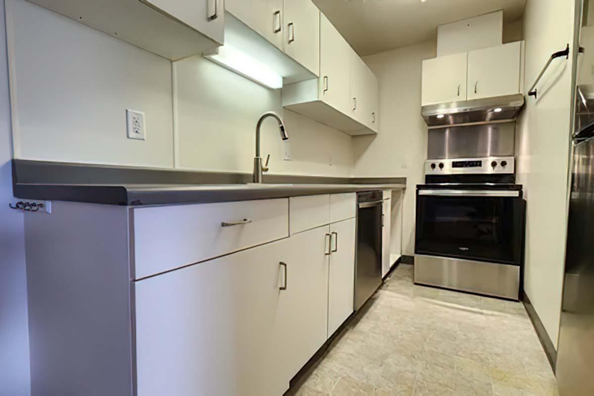 A modern kitchen featuring sleek white cabinetry, a stainless steel sink, a dishwasher, and an oven range. The countertop is gray, and the flooring appears light-colored. There is under-cabinet lighting above the countertop, creating a bright and inviting space.
