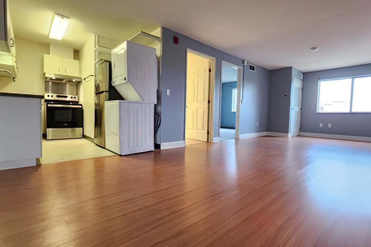 A spacious living area with hardwood flooring and a small kitchen in the background. The kitchen features modern appliances, including a stove and refrigerator. There are two doorways leading to other rooms, and a large window allowing natural light to enter the space. The walls are painted a soft blue.