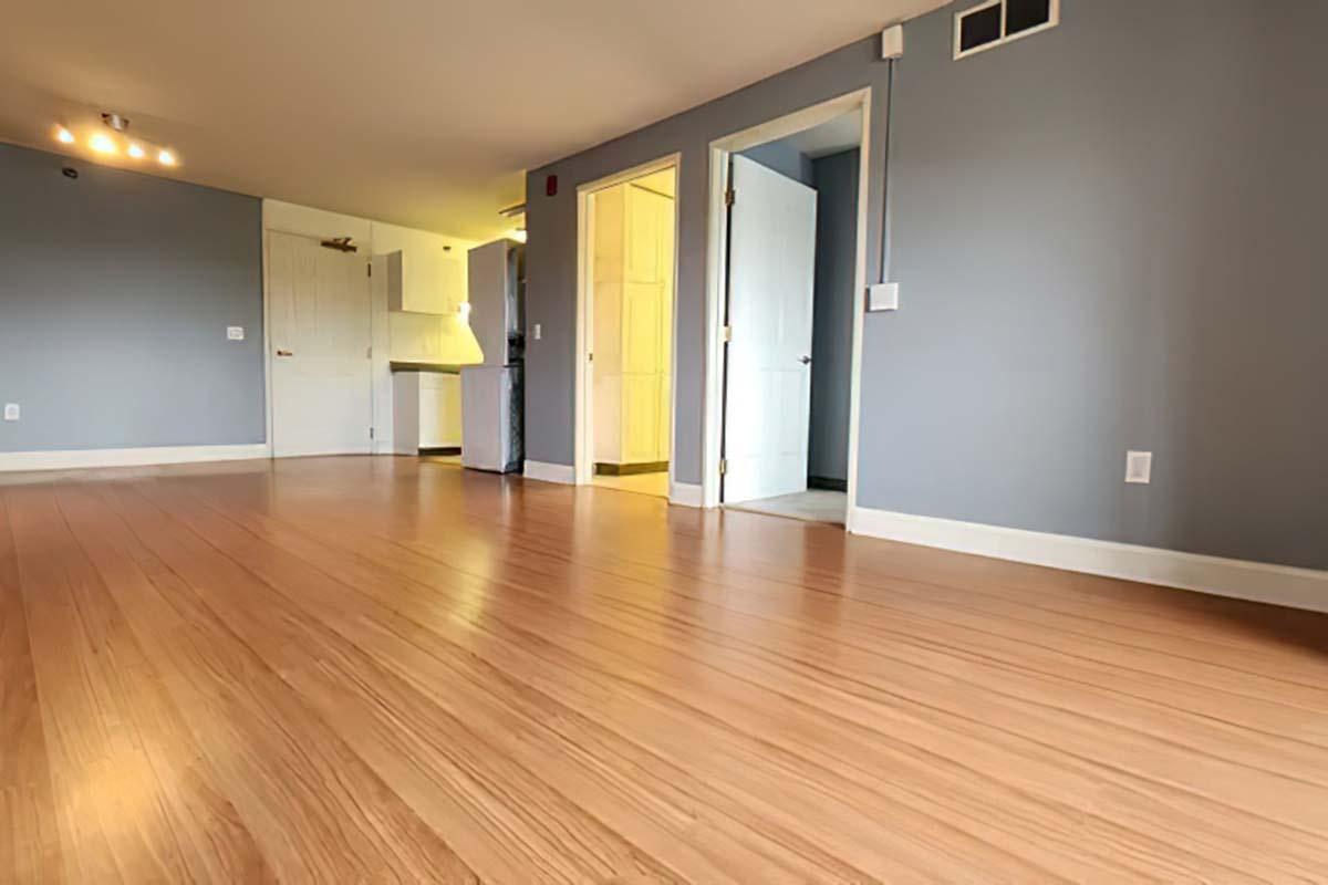 A spacious room with light brown hardwood flooring, gray walls, and an open doorway leading to another area. There are two doors visible: one open and another closed. The room is well-lit with natural light coming from a nearby window, creating a bright, inviting atmosphere.