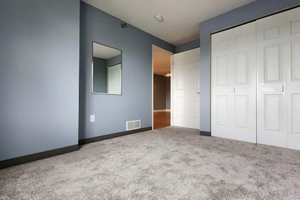 A spacious room with light gray walls and plush carpet. There is a small mirror mounted on one wall, and a doorway leading to another room can be seen. The closet features double white doors, and the overall lighting is bright and inviting.