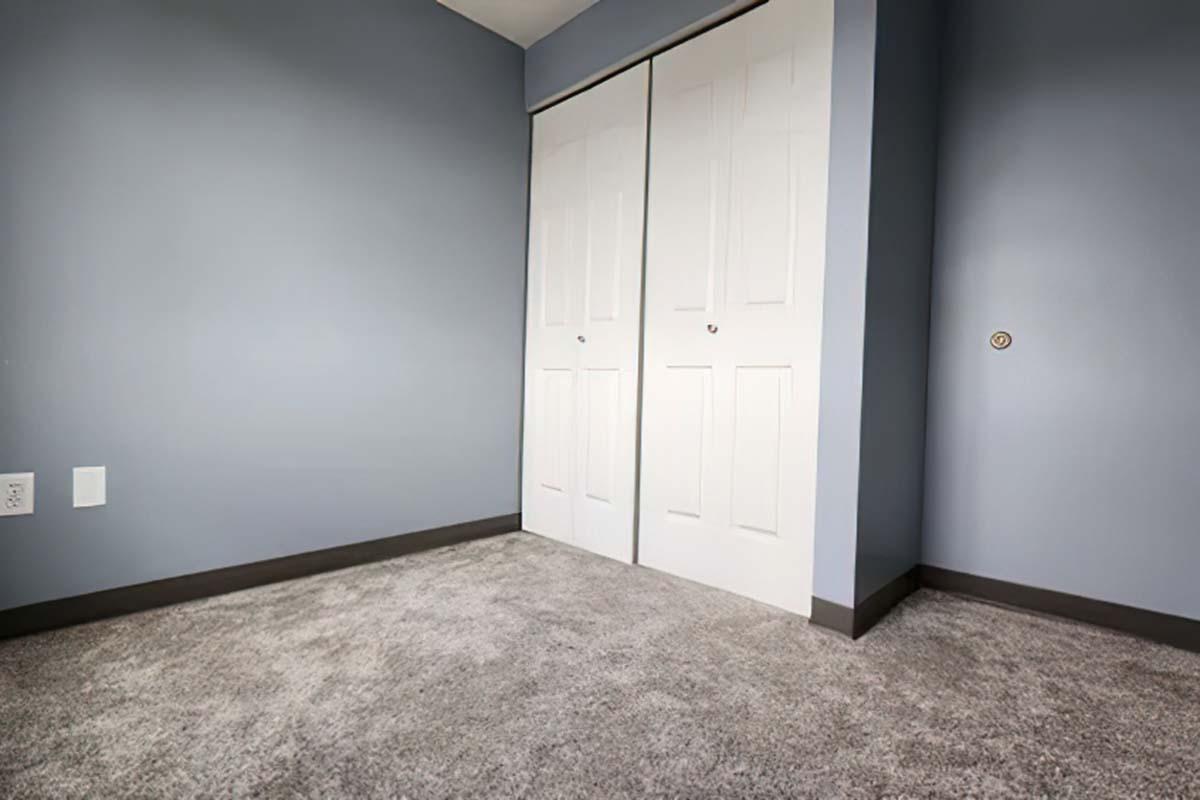 A small, empty room with light blue walls and gray carpeting. There are double white doors for a closet on the left side. The lighting is soft, and there is a power outlet visible on the left wall. The overall ambiance is calm and minimalistic.