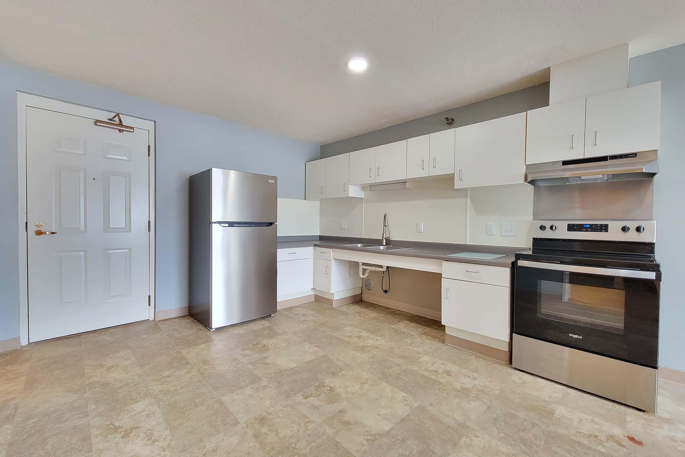 Modern kitchen featuring a stainless steel refrigerator, oven, and sleek cabinetry. The space has a light-colored tile floor and a neutral wall color, with a door leading to another room. Bright overhead lighting illuminates the area, creating a spacious and inviting atmosphere.
