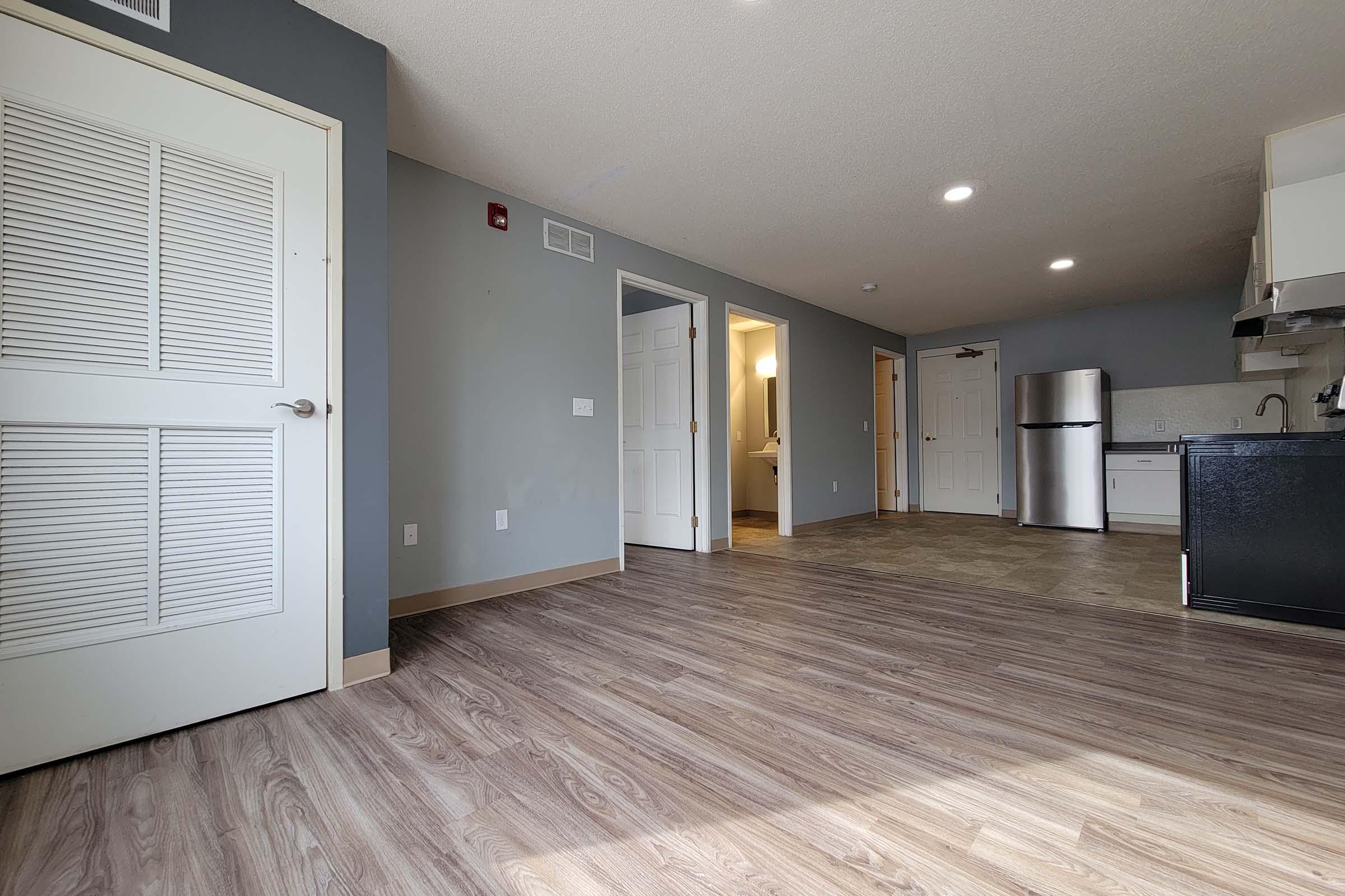 A spacious interior of an apartment featuring a living area with wood-like flooring, a kitchen area with stainless steel appliances, and multiple doorways leading to other rooms. The walls are painted in light gray and there are ample lights illuminating the space.