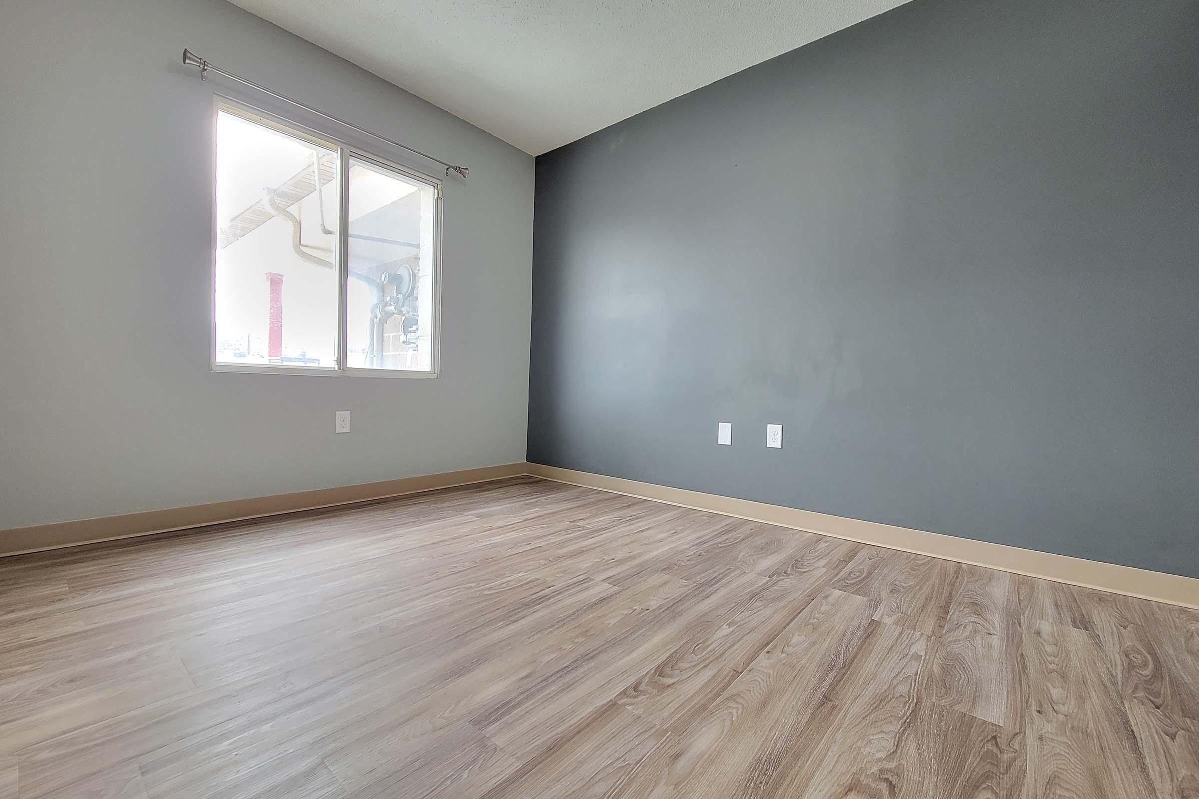 A sparsely furnished room featuring light-colored wooden flooring, a gray accent wall, and a large window allowing natural light to enter. The room appears clean and empty, with no furniture or decorations present.