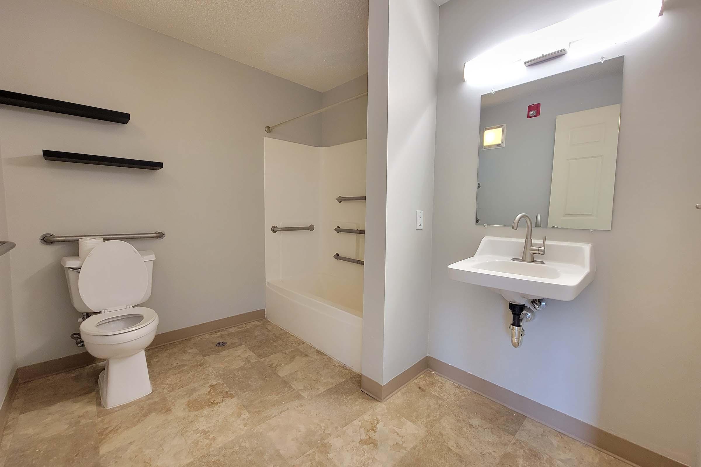 A bathroom featuring a toilet, a sink with a wall-mounted mirror, and a bathtub with grab bars. The walls are painted light gray, and there are small black shelves on one side. The floor is tiled with a light stone pattern, and natural light enters through a small window.