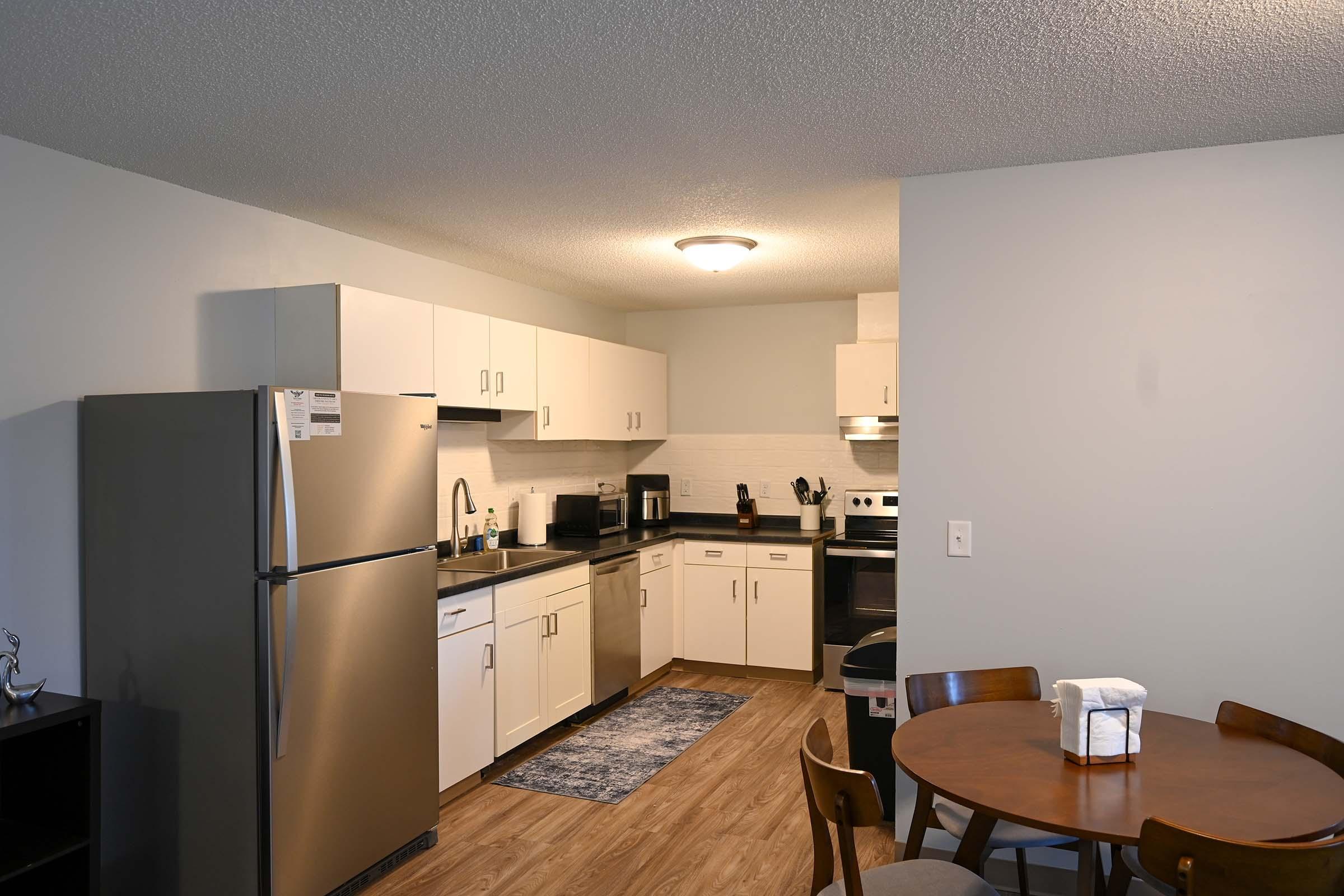 A modern kitchen featuring stainless steel appliances, including a refrigerator, microwave, and stove. The cabinetry is white with a sleek design, and there's a dark countertop. A dining table with four chairs is visible in the foreground, and a rug is placed on the floor near the kitchen area. The space is well-lit and contemporary.