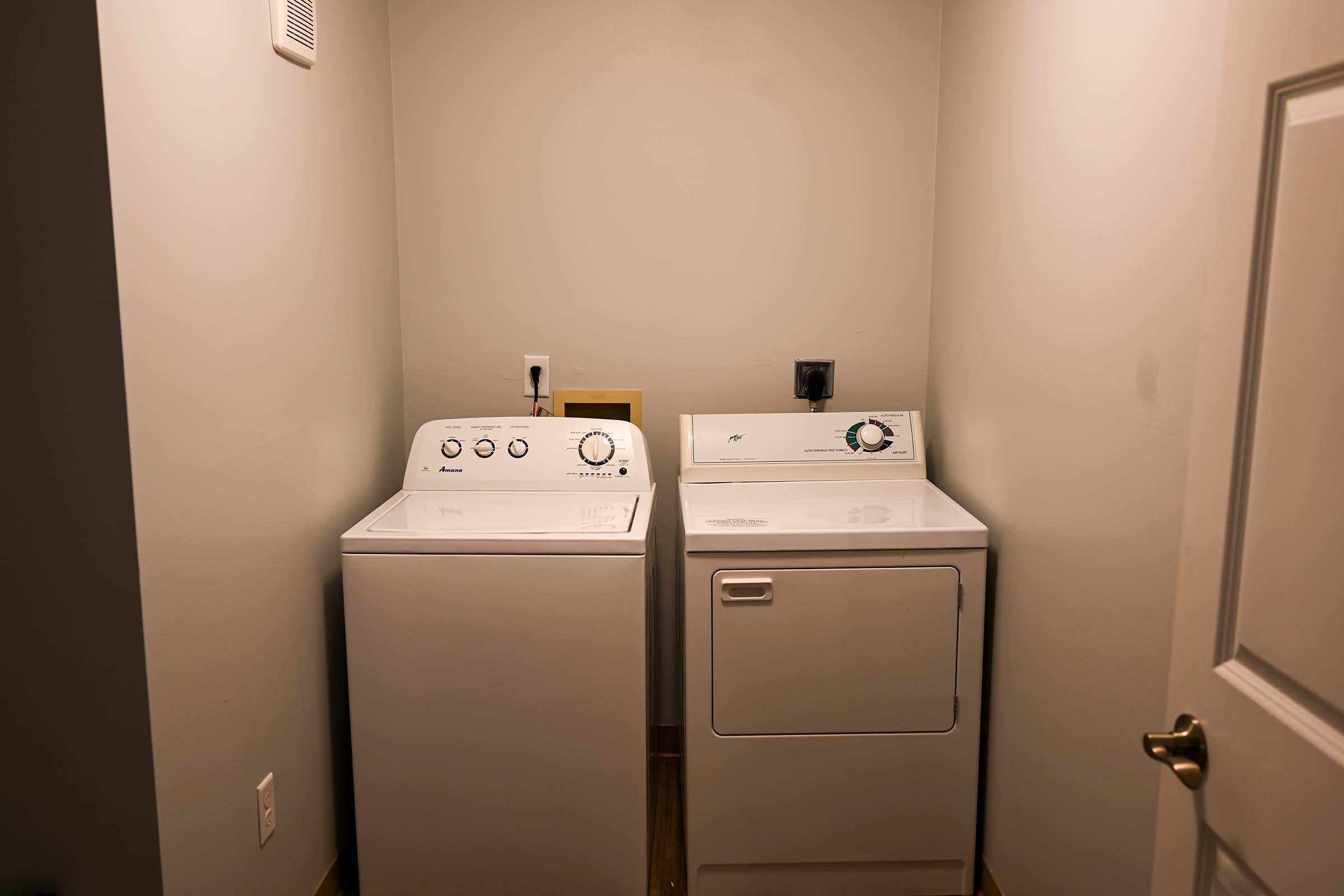 A laundry room featuring a white washing machine and a white dryer side by side against a light gray wall. The floor is hardwood, and there is a door leading out of the room. There is a power outlet on the wall beside the appliances.