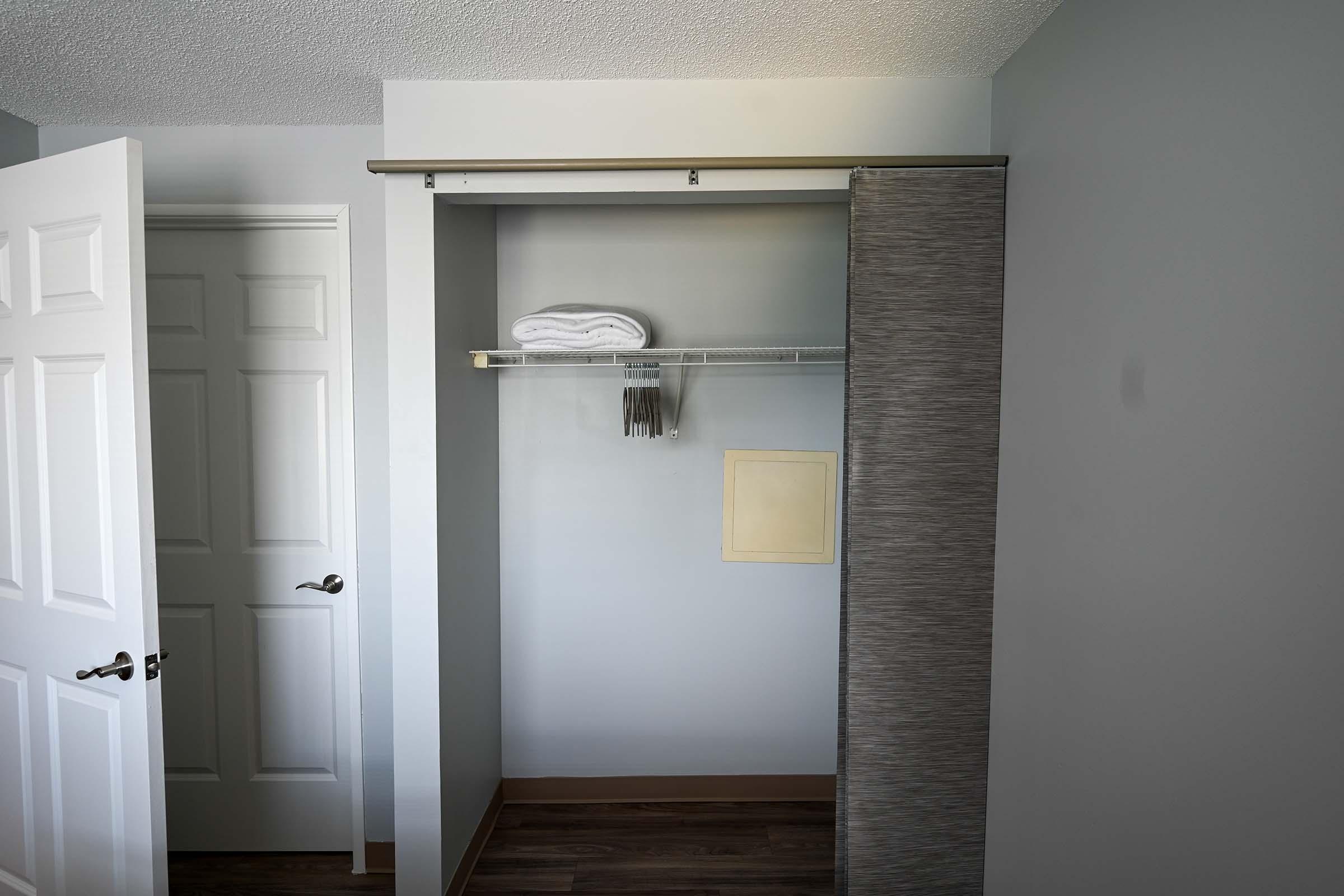 A partially empty closet with a shelf holding neatly folded towels. A wire rack is attached above the shelf. The interior walls are painted gray, and there is a small, blank space on one wall. A white door leading to another room is visible on the left. The flooring is wooden.