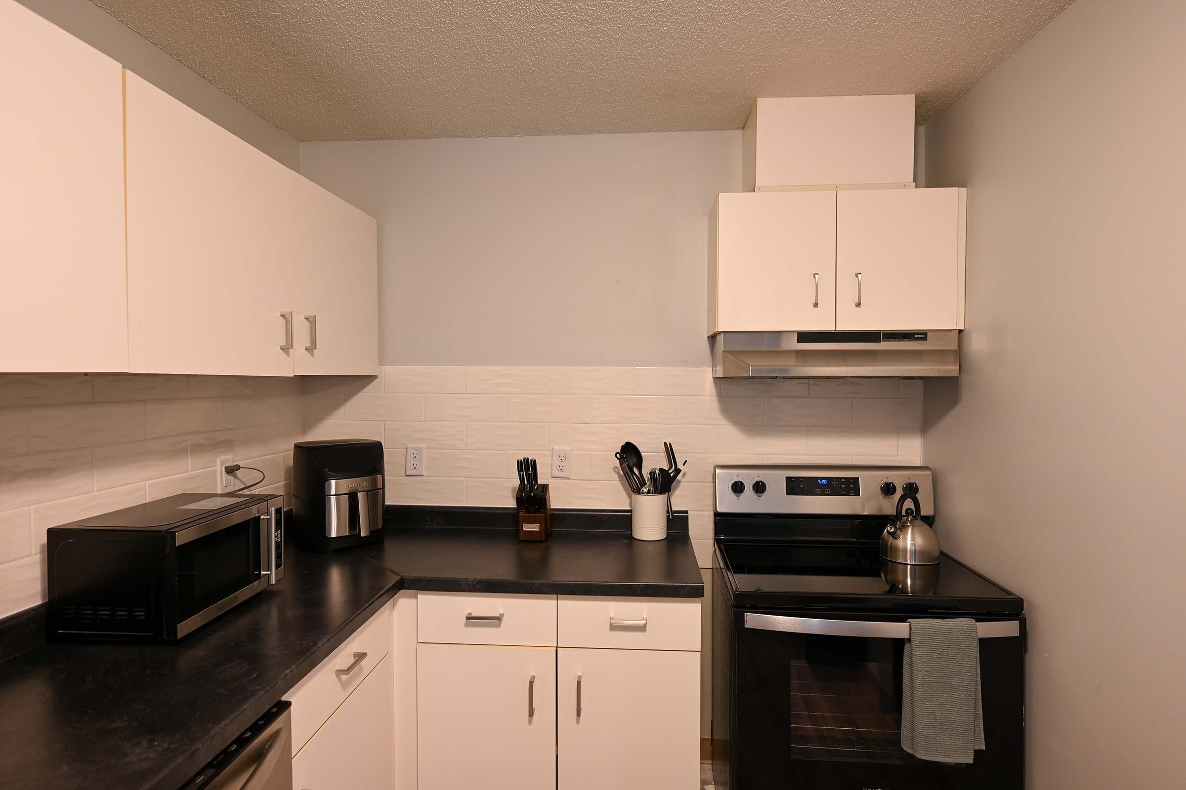 A modern kitchen featuring white cabinets, a black countertop, a stainless steel oven, and a stovetop with a kettle. There's a microwave and a coffee maker on the counter, along with kitchen utensils in a holder. The walls are painted light gray, and the atmosphere is clean and organized.