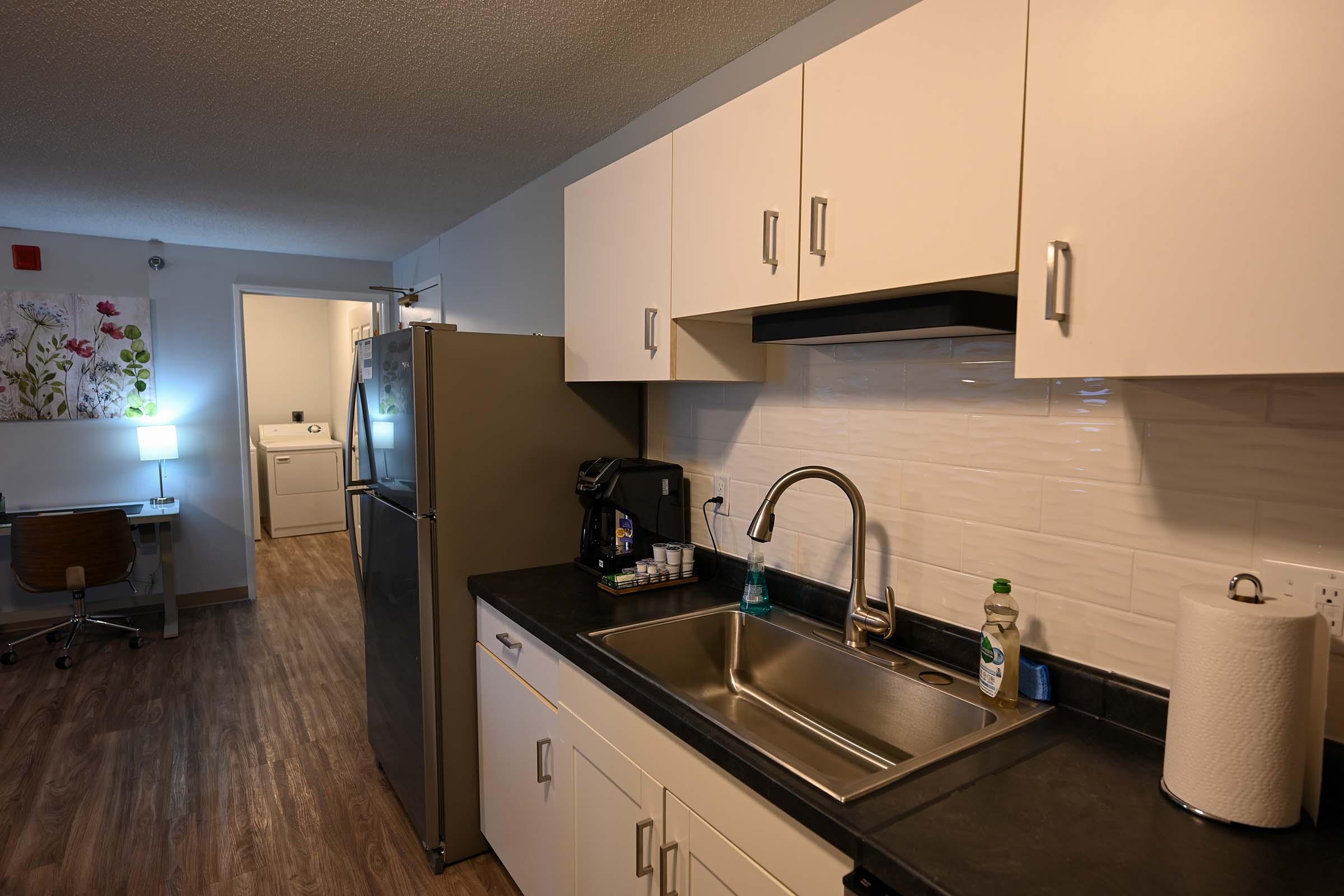 A modern kitchenette featuring a stainless steel sink, a gray refrigerator, and a coffee maker on the counter. White cabinets above the sink complement the dark countertop. In the background, a clean workspace with a desk and a laundry area can be seen. Natural light enters from a nearby room.