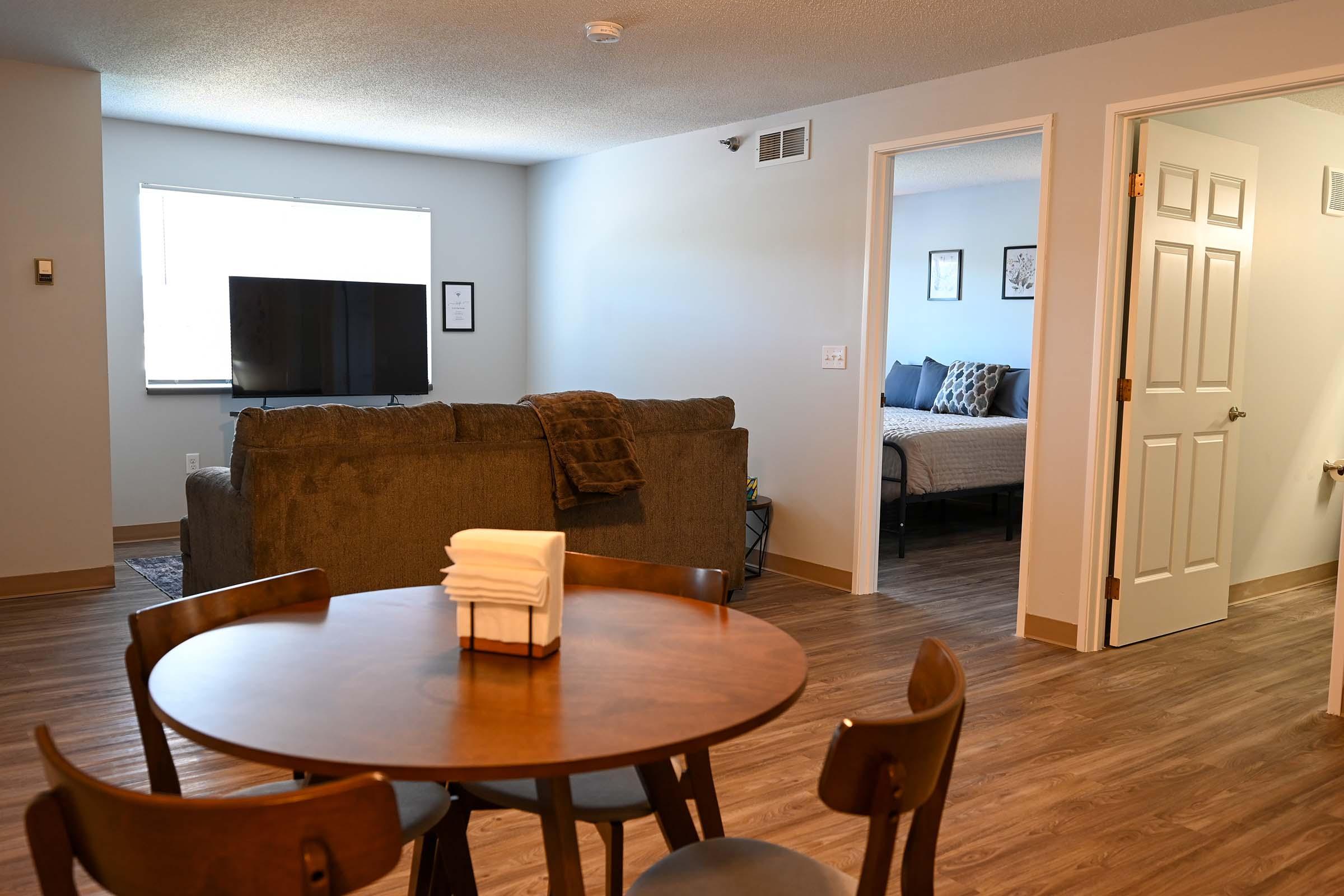 A modern living space featuring a brown couch and a flat-screen TV in the foreground, with a round wooden dining table and neatly arranged towels. In the background, a doorway leads to a bedroom with a bed and decor visible. The room has light-colored walls and a window allowing natural light.