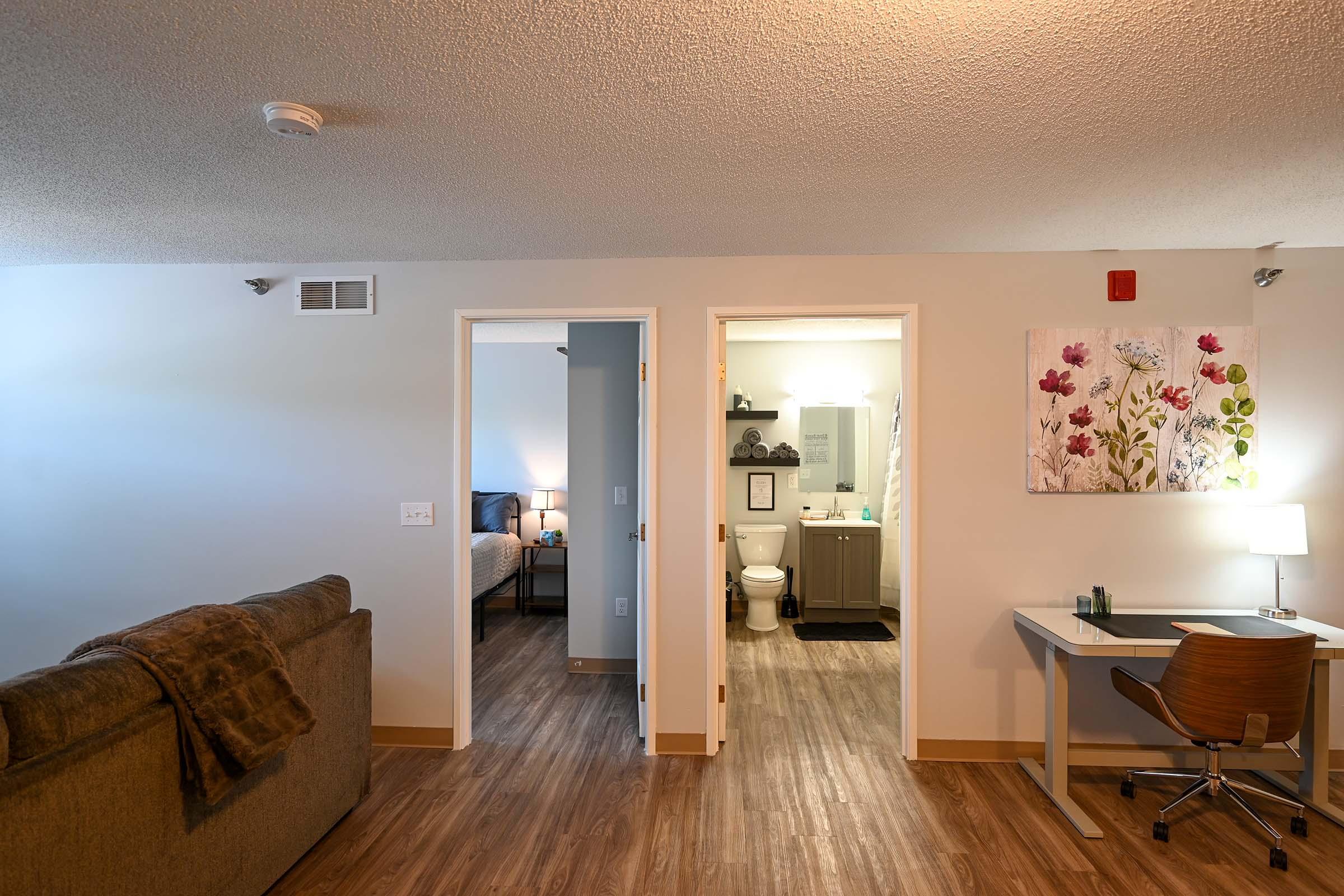 A view of a modern apartment interior, featuring a cozy living area with a sofa, a desk, and an open doorway leading to a bathroom and a bedroom. The bathroom is visible with a toilet and sink, while floral wall art adds a touch of decor to the space. Warm lighting creates an inviting atmosphere.