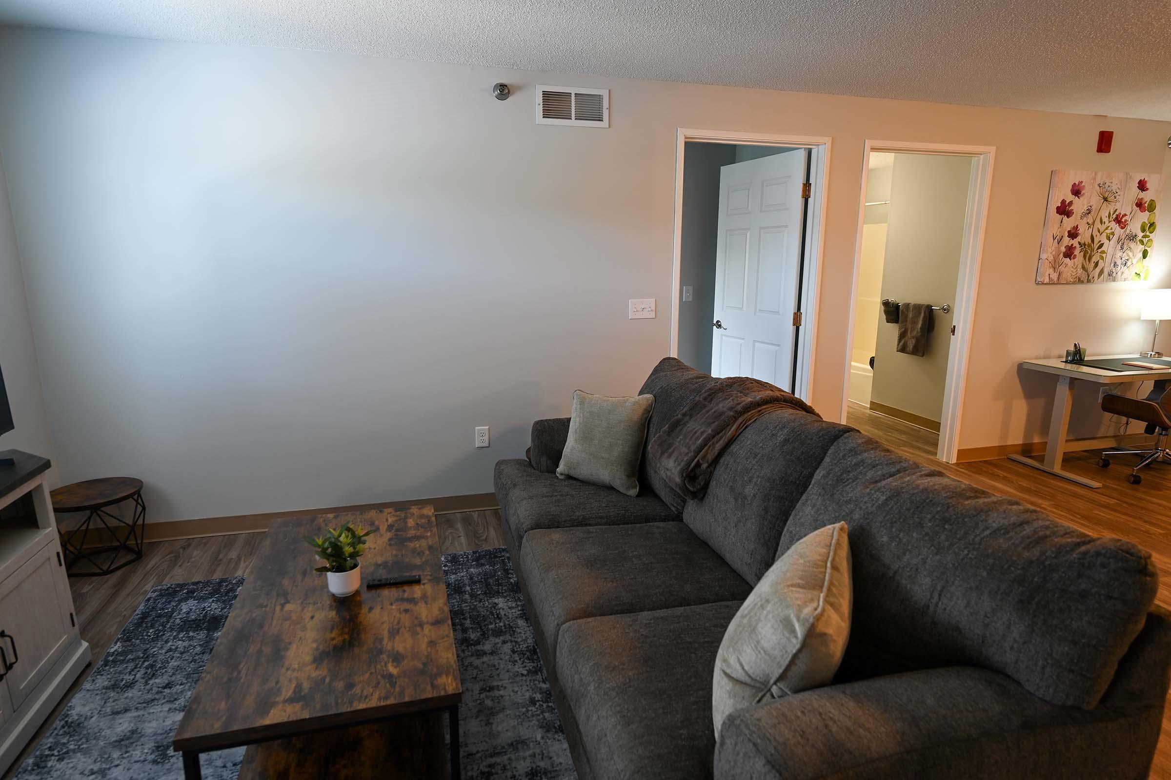 A cozy living room featuring a gray sectional sofa adorned with a decorative pillow, a wooden coffee table, and a small potted plant. In the background, a partially open white door leads to a bathroom, while a desk and chair are visible on the right, illuminated by soft lighting. The walls are painted in neutral tones.