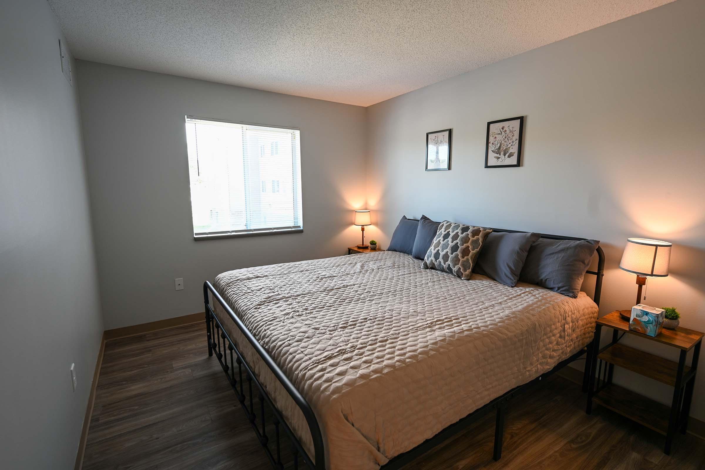A cozy bedroom featuring a large bed with gray bedding and decorative pillows. There's a window allowing natural light, two small bedside tables with lamps, and framed wall art. The walls and floor have neutral tones, creating a warm and inviting atmosphere.