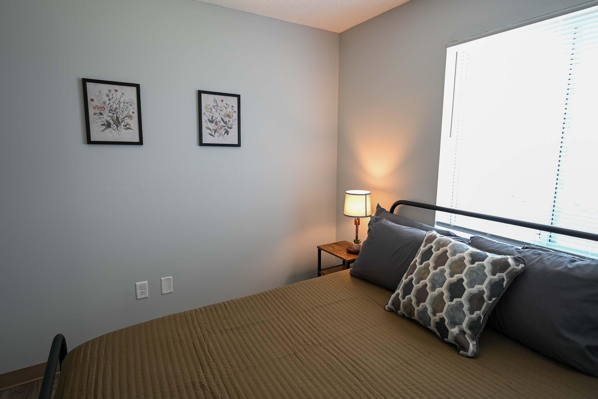 A cozy bedroom featuring a bed with a tan quilt and decorative pillows. Two framed floral prints hang on the wall beside a window. A bedside table supports a warm lamp, creating a welcoming atmosphere with soft lighting. The walls are painted in a light gray color.