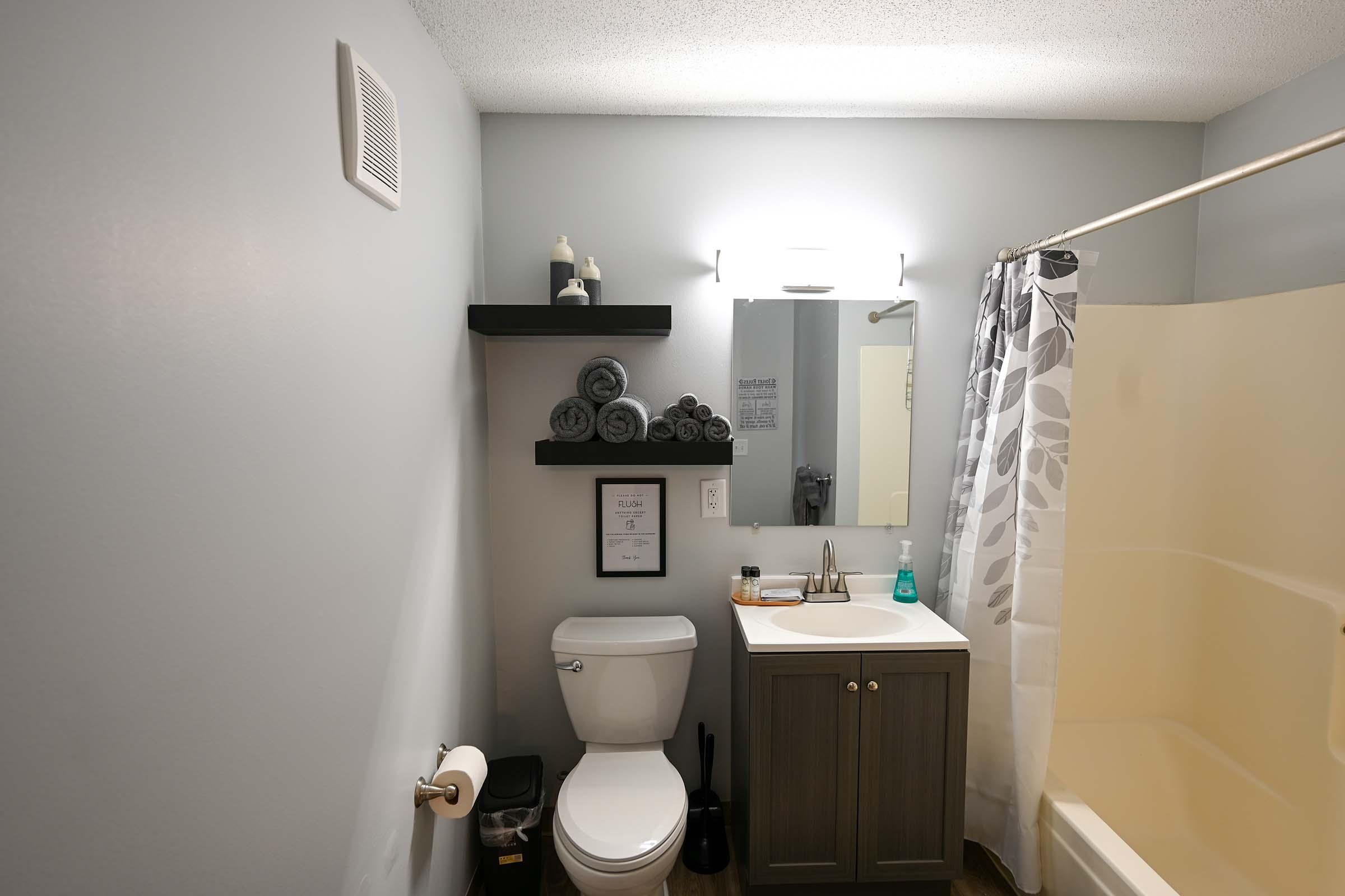 A modern bathroom featuring a toilet, sink, and mirror. There are neatly stacked gray towels on a black shelf above the sink. A shower curtain with a floral design is visible, along with a bathtub. Accessories like soap and a hand sanitizer are placed nearby, and the walls are painted a soft gray.