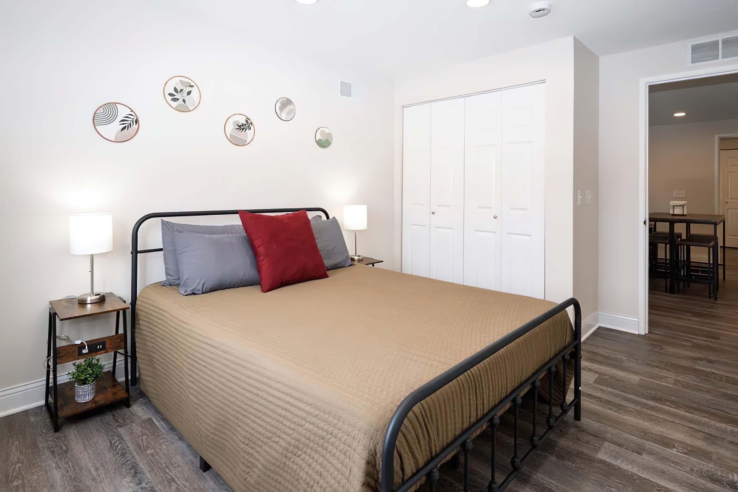 A cozy bedroom featuring a metal bed frame with a tan quilt and gray pillows, accompanied by a red accent pillow. There are two bedside tables with lamps, decorative wall plates, and a closet in the background. The flooring is wooden, and the walls are painted light.