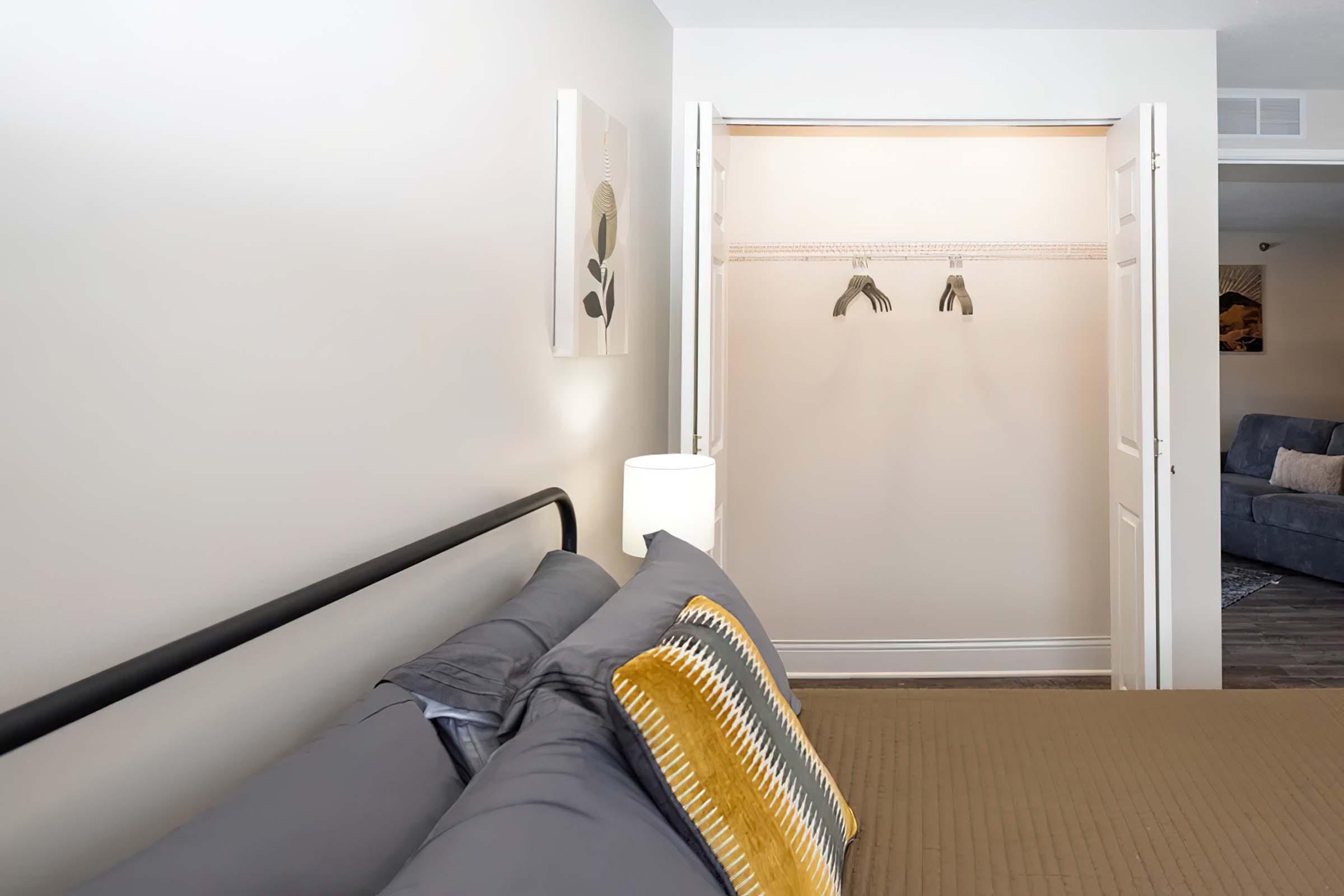 A cozy bedroom featuring a bed with gray and yellow pillows, a bedside lamp, and a closet with open doors revealing two hangers. The walls are painted in a soft color, and there's a framed artwork on one wall. The floor has a warm, wooden appearance, giving a welcoming atmosphere.