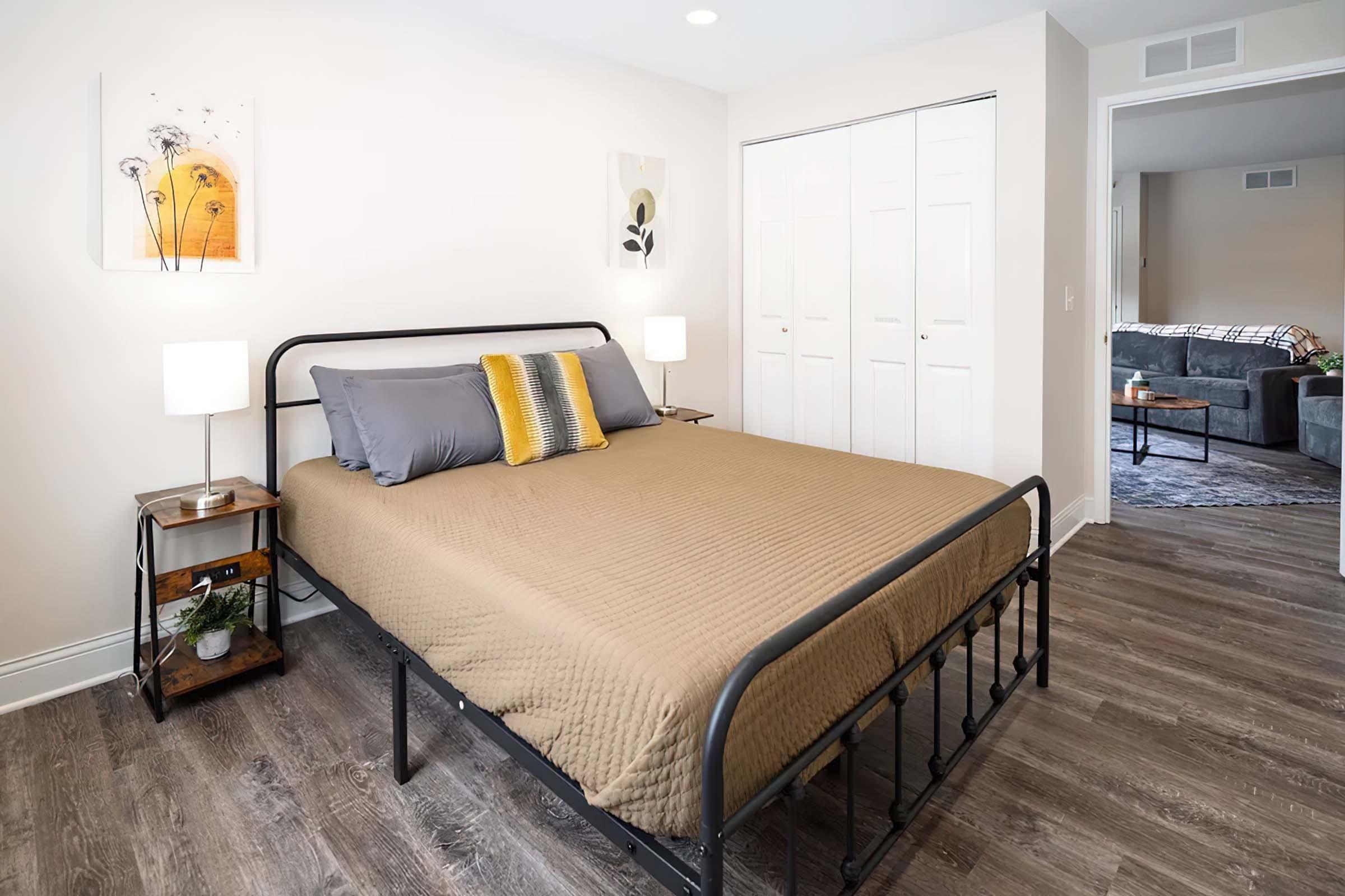 A cozy bedroom featuring a neatly made bed with gray and yellow bedding, a nightstand with a lamp, and artwork on the wall. There are sliding closet doors and a glimpse of a living area visible in the background, which has a couch and additional decor. The room is well-lit and inviting.