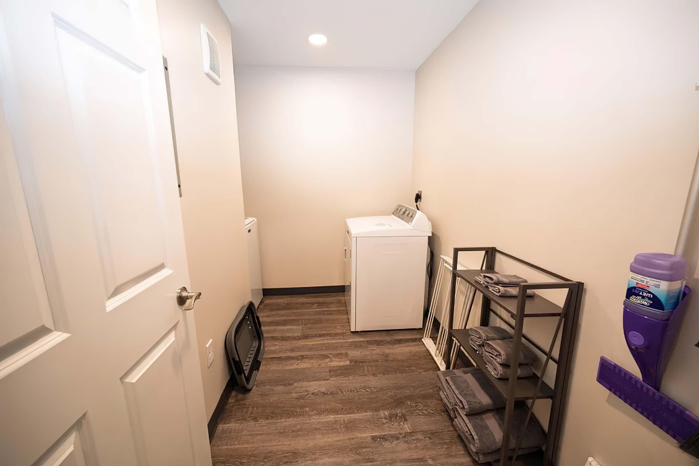 A small laundry room featuring a white washing machine, a shelving unit with neatly stacked towels, and a wall-mounted storage holder with cleaning supplies. The walls are painted light beige, and the floor is dark wood-like laminate. There is a door on the left side and soft lighting in the ceiling.