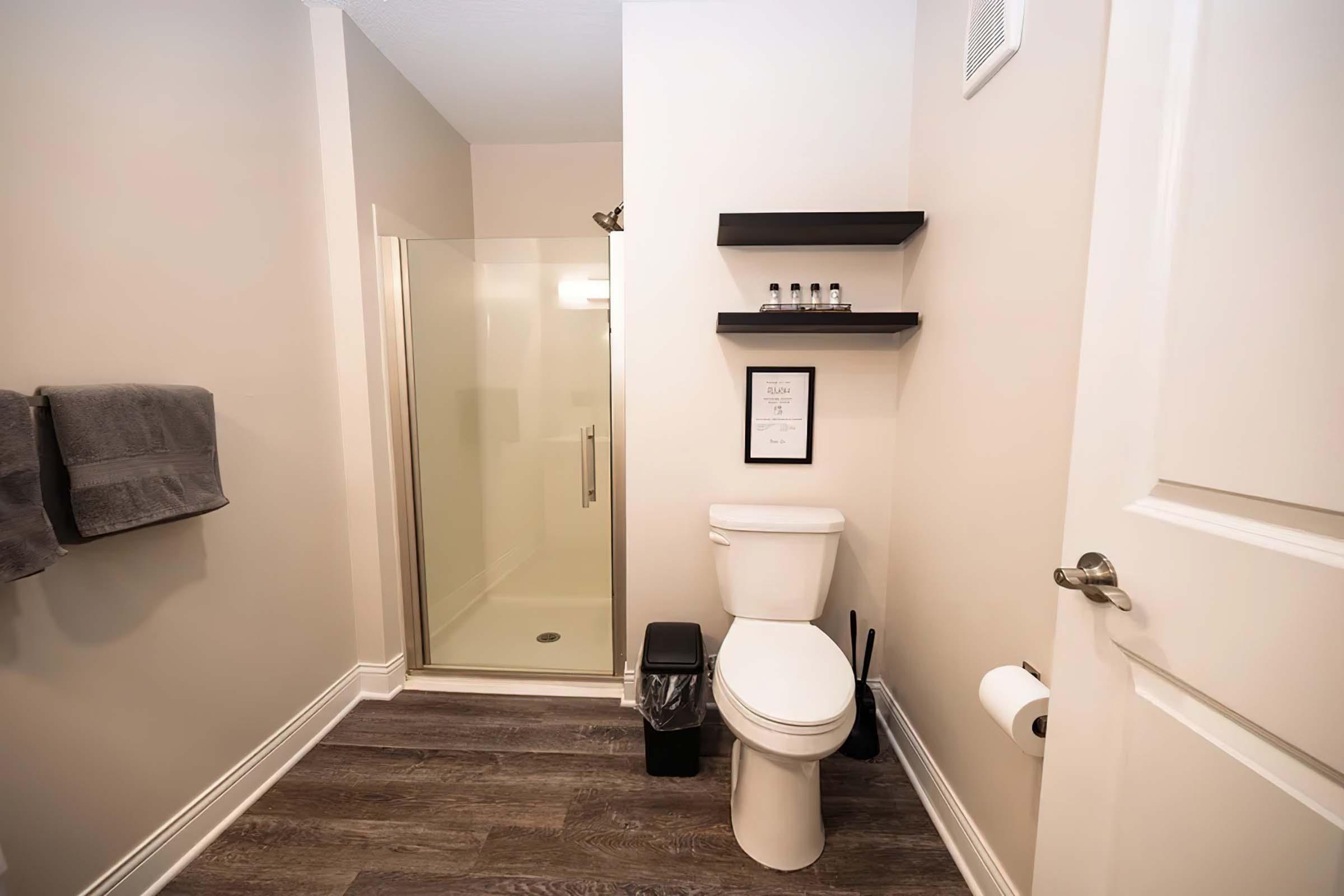 A clean, modern bathroom featuring a toilet, a shower stall with a glass door, a small shelf with toiletries, and neatly arranged towels. The walls are painted in a light color, and there is a trash can and toilet paper within easy reach. The flooring is dark wood laminate.